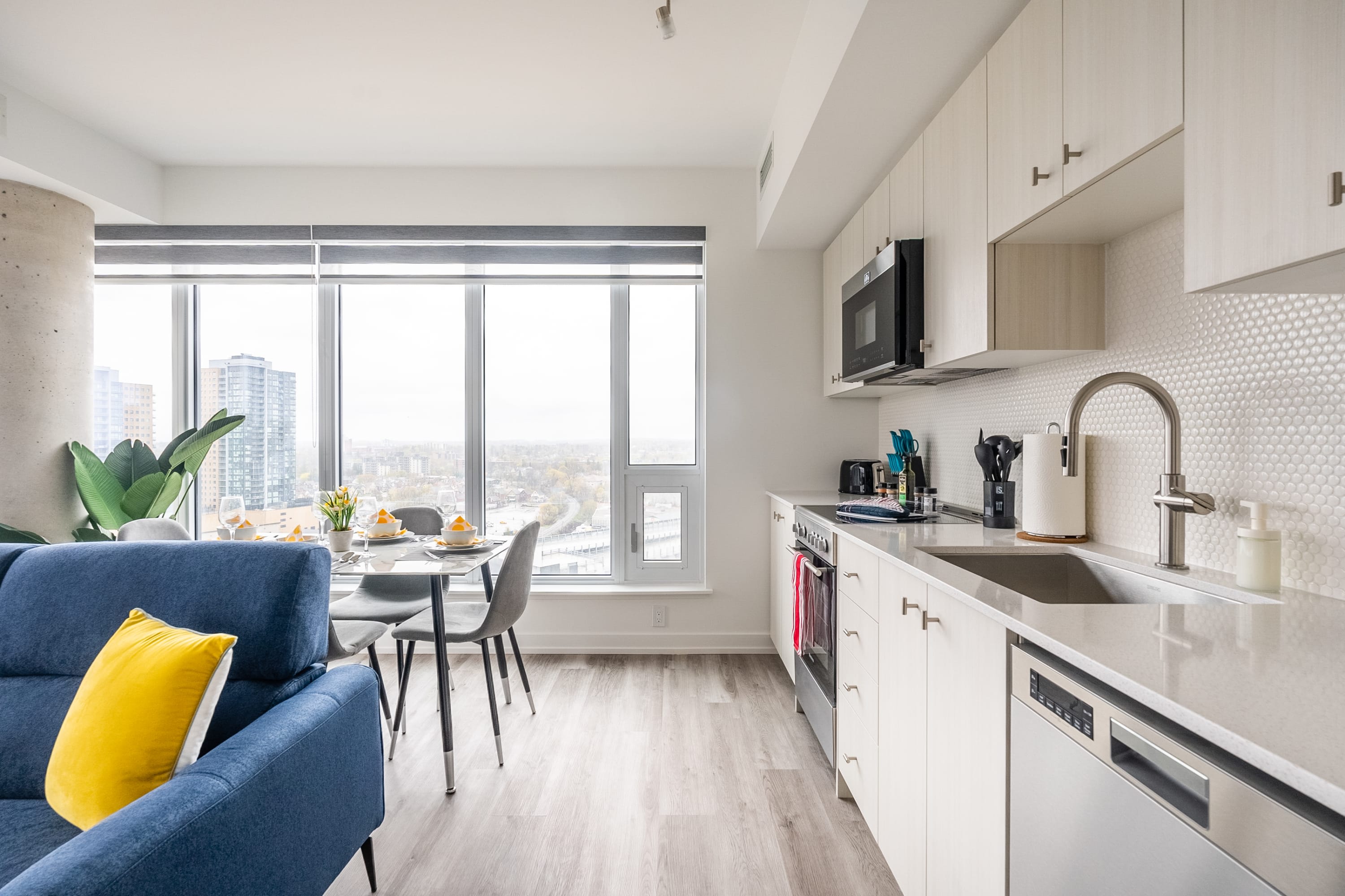 Modern kitchen with light wood upper and lower cabinets, white subway tile backsplash, stainless steel appliances including a microwave, stove, and dishwasher, and a large sink, adjacent to a dining area and large windows.