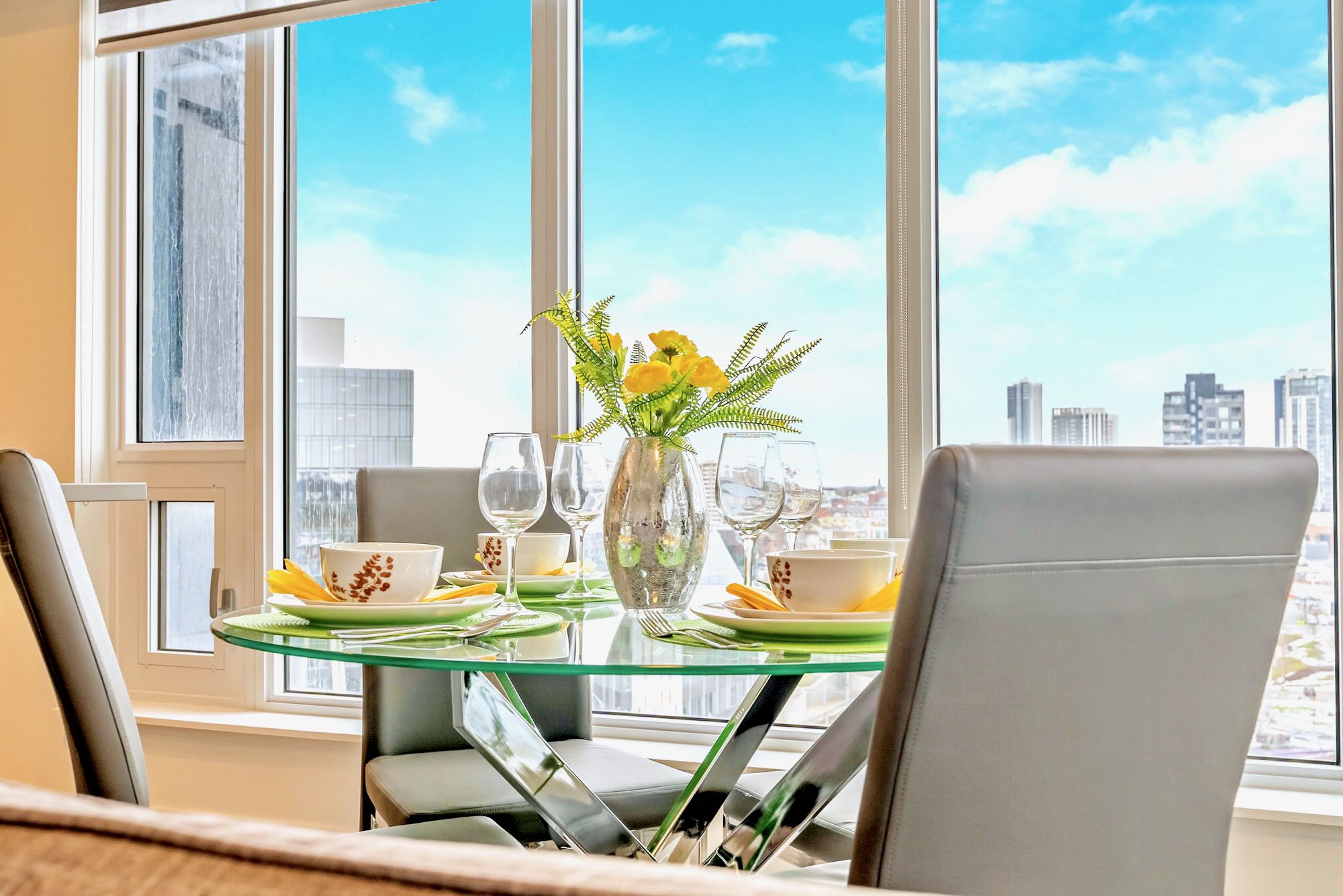 Close-up of a modern dining table set for four, featuring a round glass top and grey upholstered chairs. A vase with yellow flowers serves as a centerpiece, with city views visible through the large window.