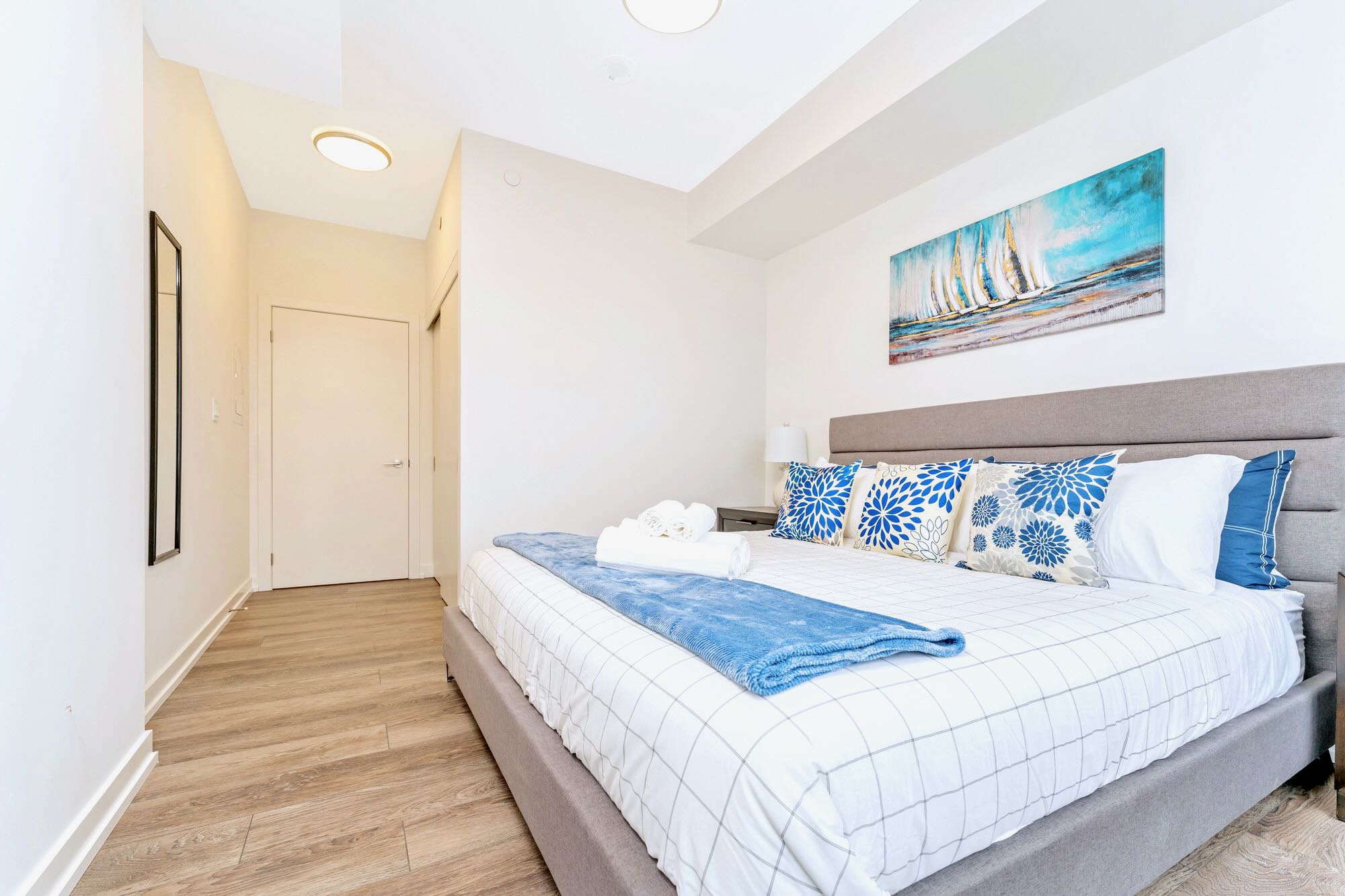 Bright bedroom with a large bed featuring a grey upholstered headboard, white and blue patterned bedding, and a blue throw, complemented by a vibrant sailboat painting above the bed and light wood flooring.
