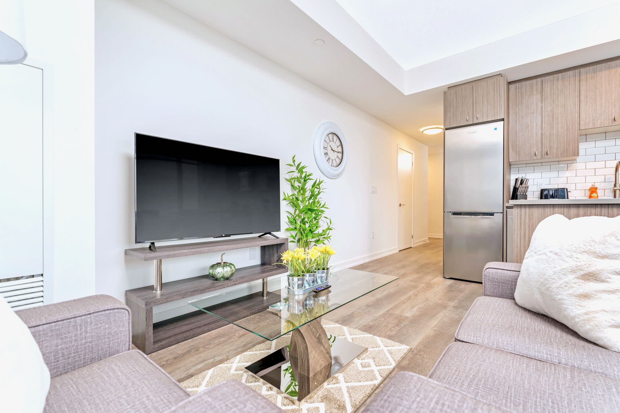 Cozy living room featuring a large flat-screen TV on a modern media console, a grey sofa, and light wood-look flooring. A large white wall clock and a potted plant add decorative touches.
