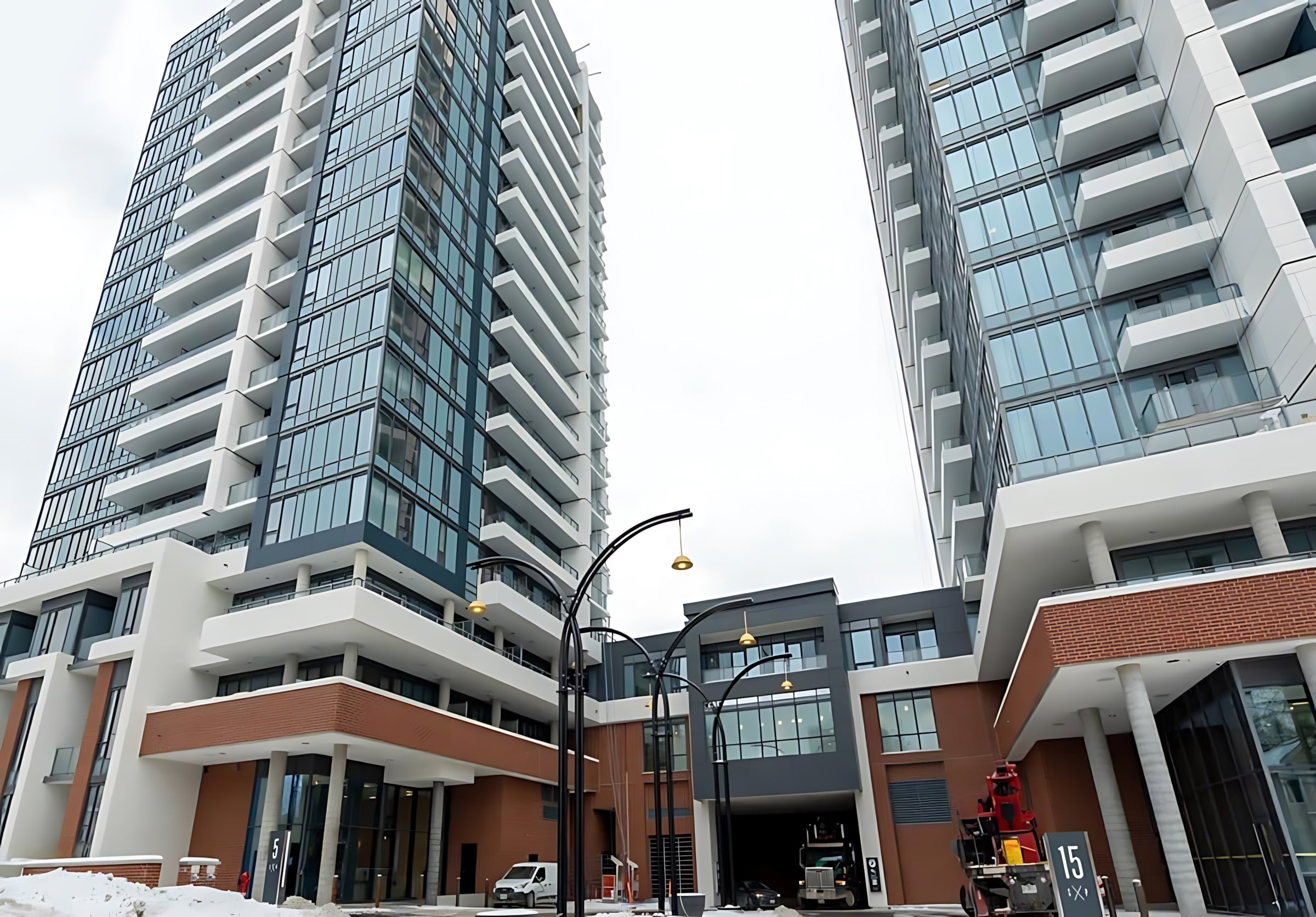 View of two modern high-rise apartment buildings with glass facades and balconies, featuring a multi-level base with brick and dark grey accents, under a cloudy sky.