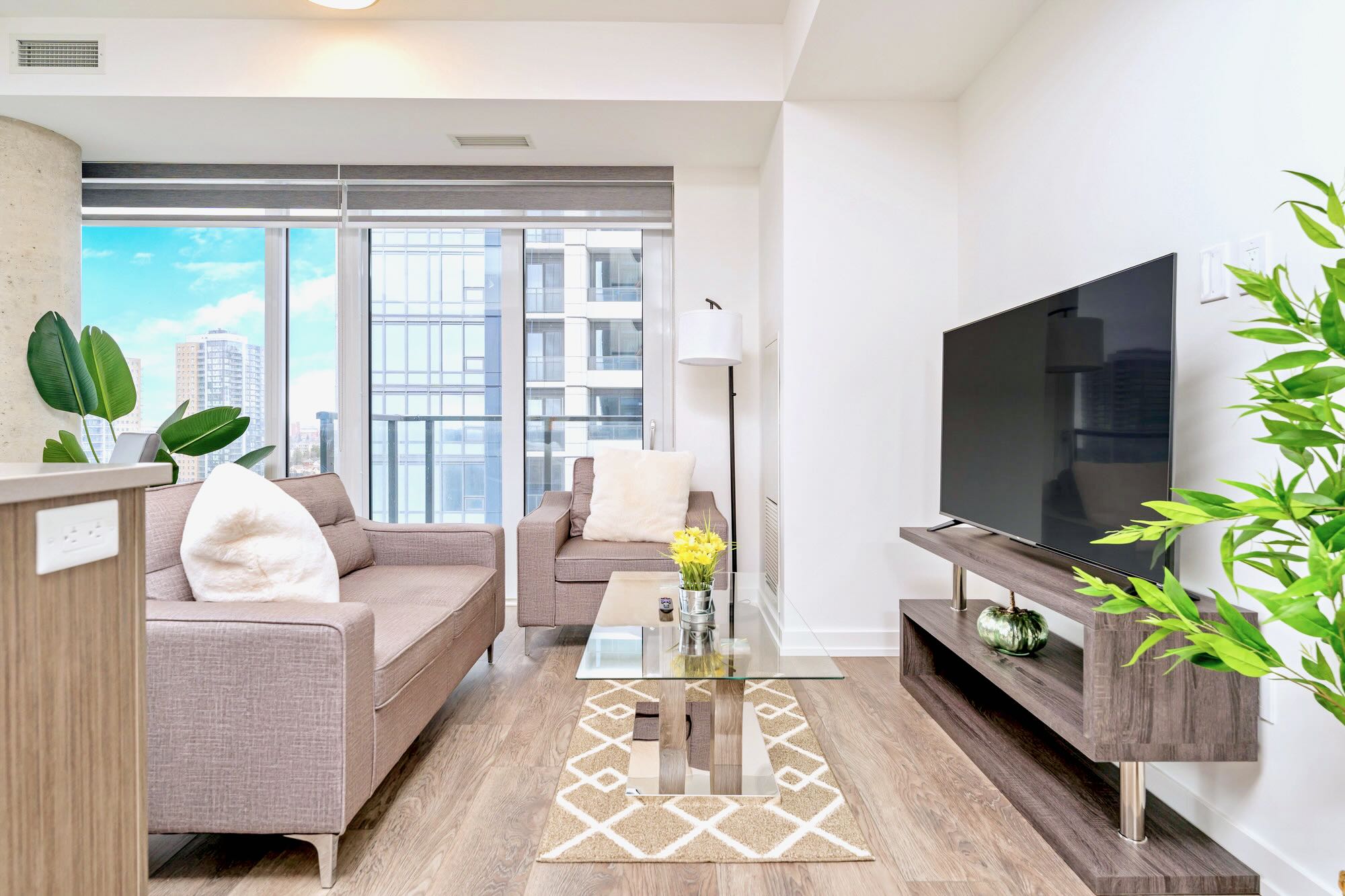 Modern living room with a large flat-screen TV on a dark wood media console, a grey sofa, and light wood-look flooring. A glass coffee table with a decorative rug sits in front of the sofa.