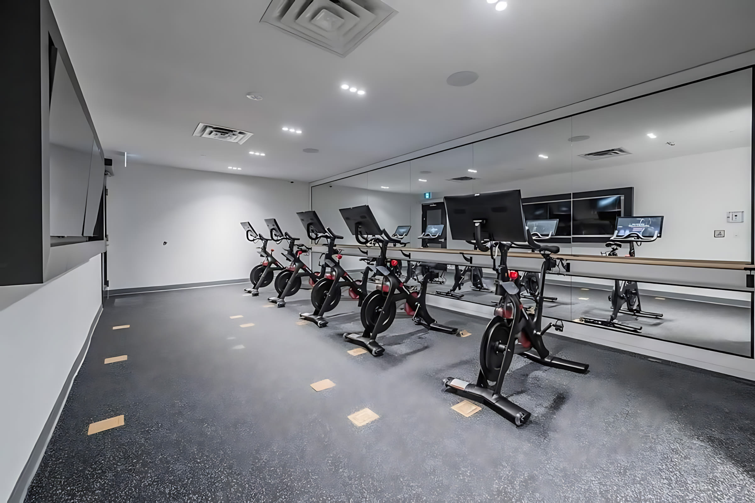 A modern fitness room features several black stationary bikes with screens, arranged in rows facing a large mirrored wall. The room has a dark rubber floor with light square accents and bright recessed lighting.