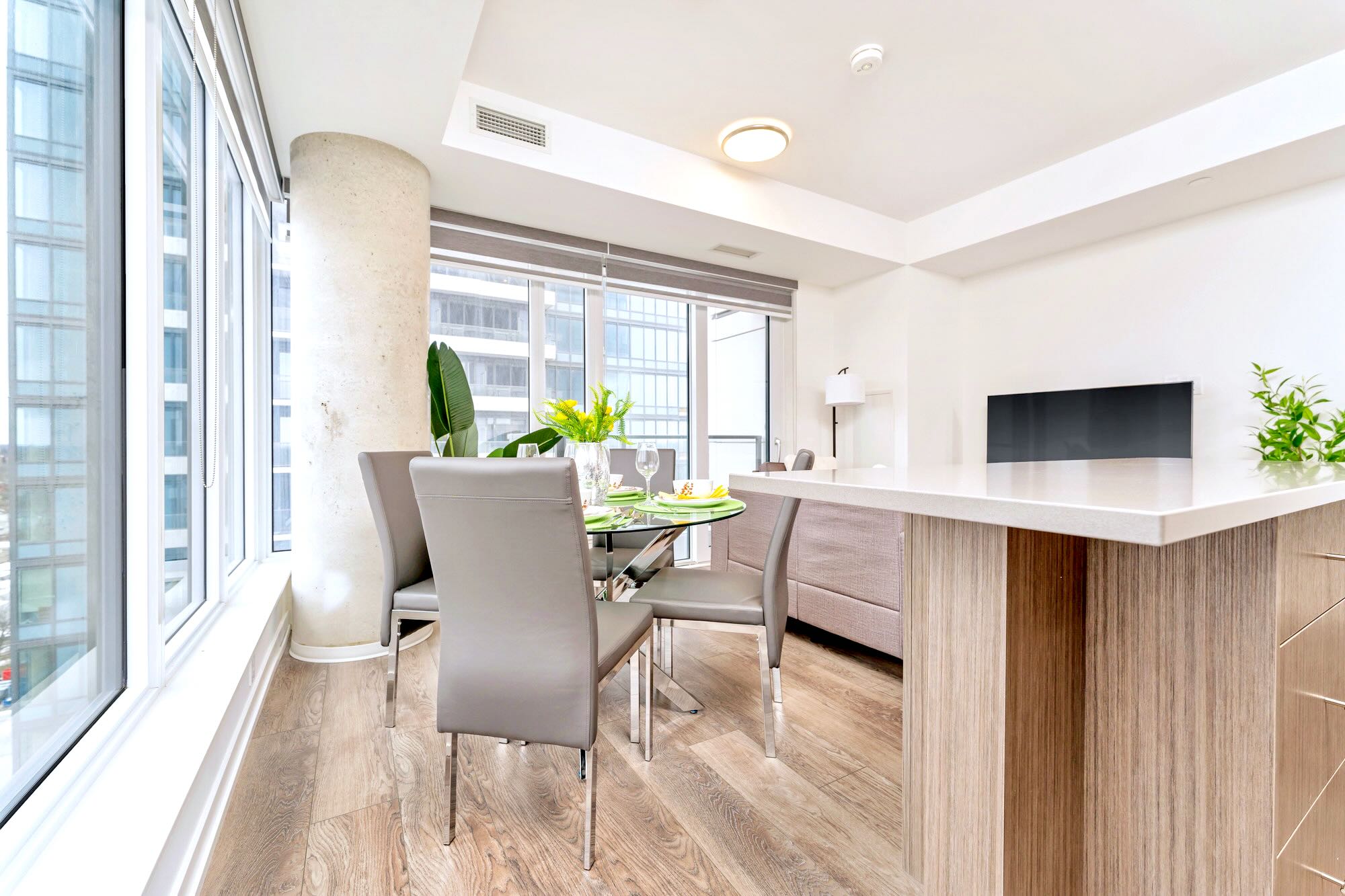 Modern dining area with a round glass table and four grey chairs, set against large windows offering city views, adjacent to a light-colored kitchen island and living space with a sofa.