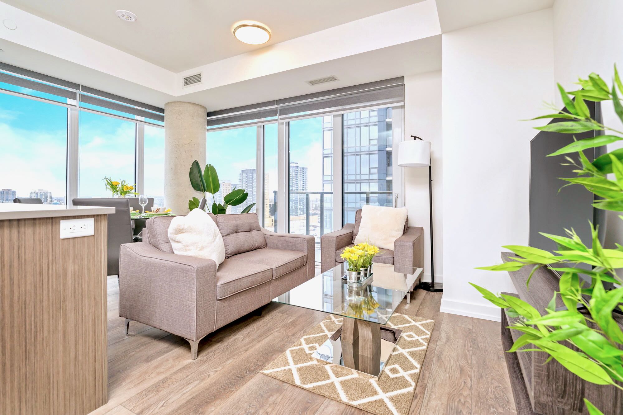 Bright living room with a grey sofa and armchair, a glass coffee table, and light wood-look flooring. Floor-to-ceiling windows offer city views, and a tall potted plant adds greenery.