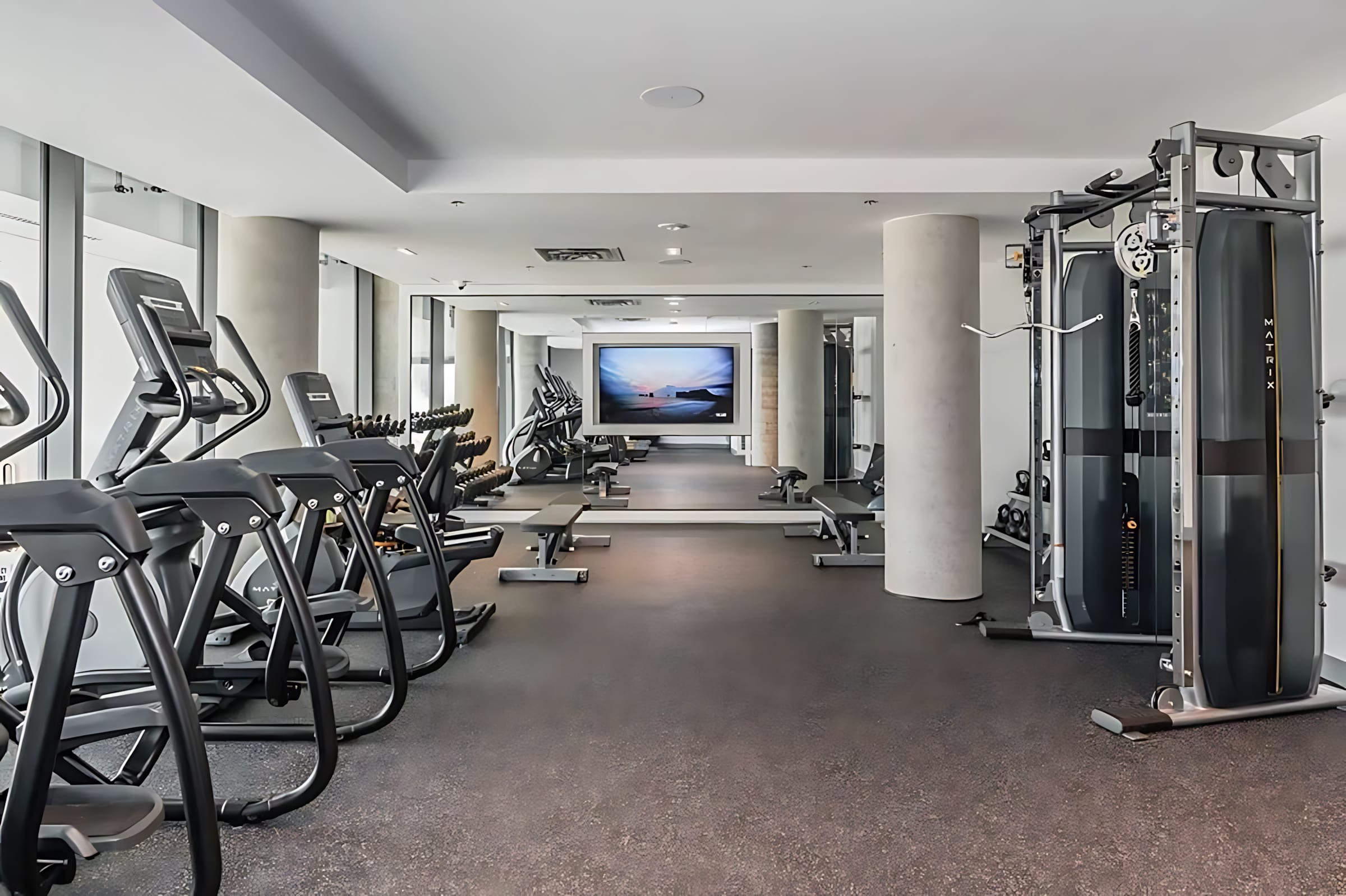 Spacious and well-equipped fitness center with various cardio machines, weightlifting equipment, and free weights, featuring a large screen displaying a scenic image at the far end.