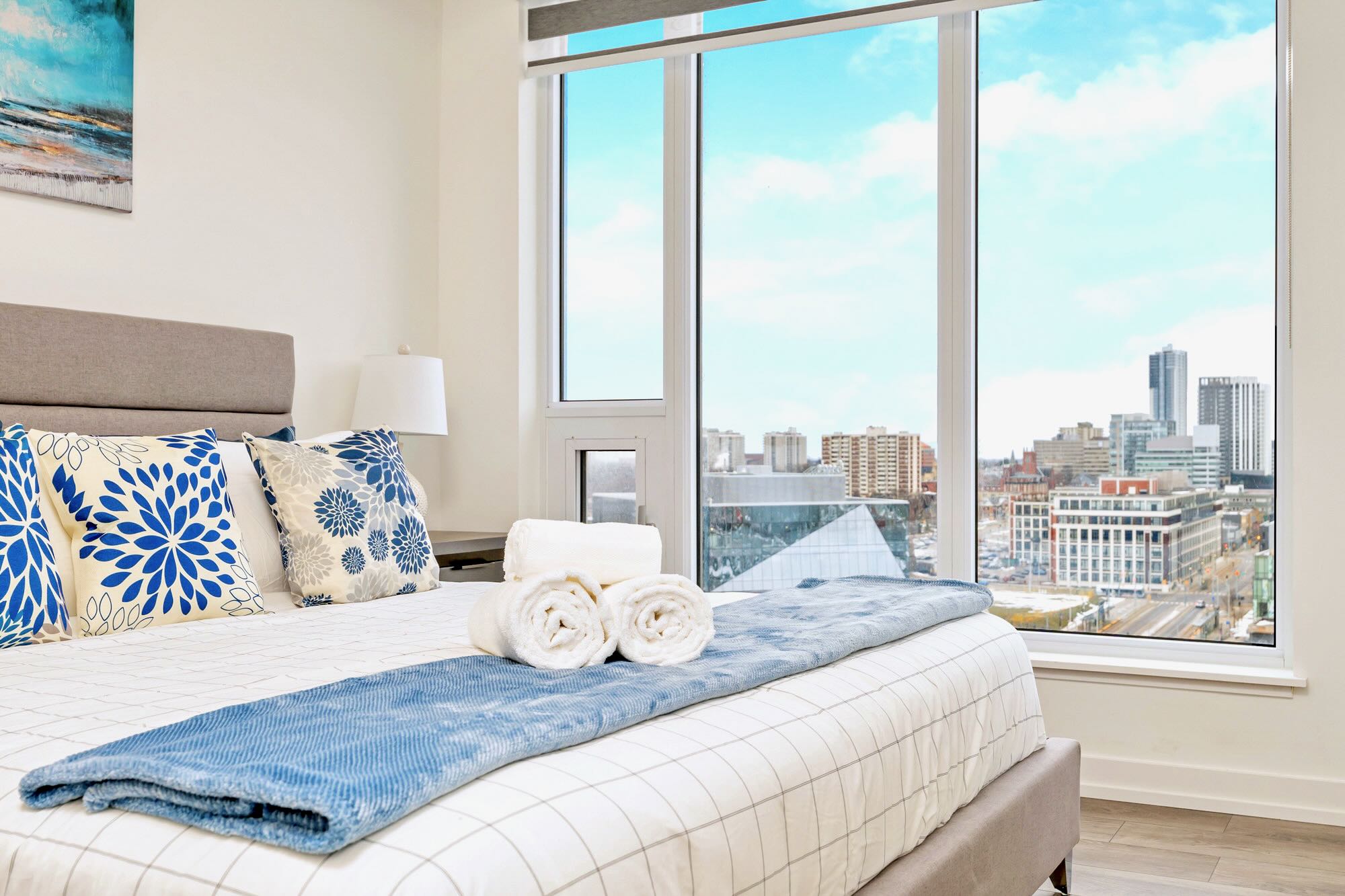 Bedroom with a large bed dressed in white and blue bedding, featuring rolled white towels, positioned next to expansive floor-to-ceiling windows that offer a breathtaking panoramic city skyline view.