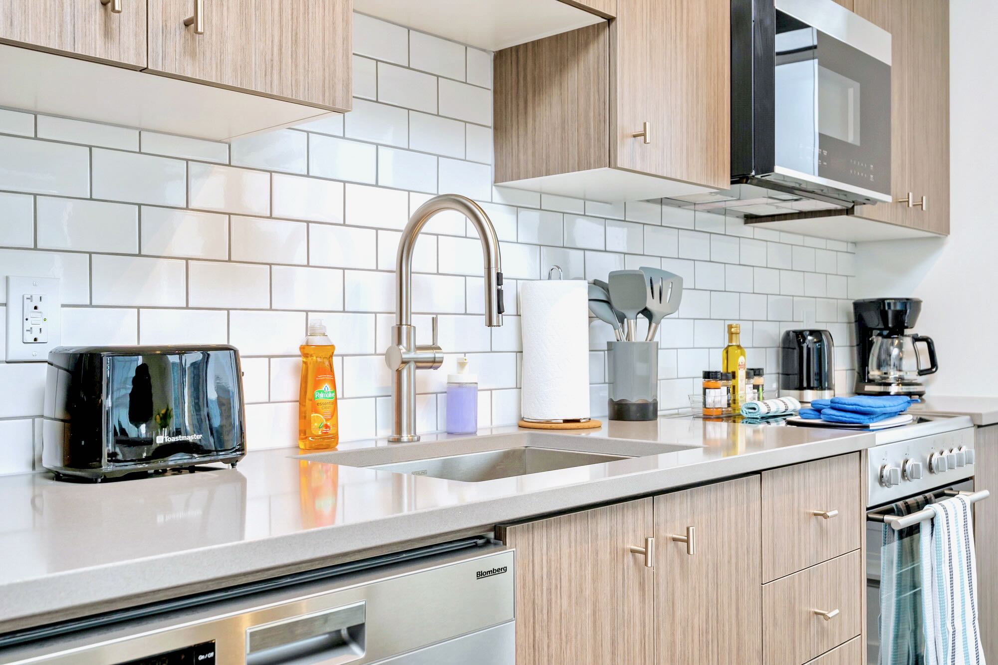 Contemporary kitchen featuring light wood cabinetry, white subway tile backsplash, stainless steel appliances including a dishwasher, sink, and microwave, with various kitchen essentials on the light grey countertop.