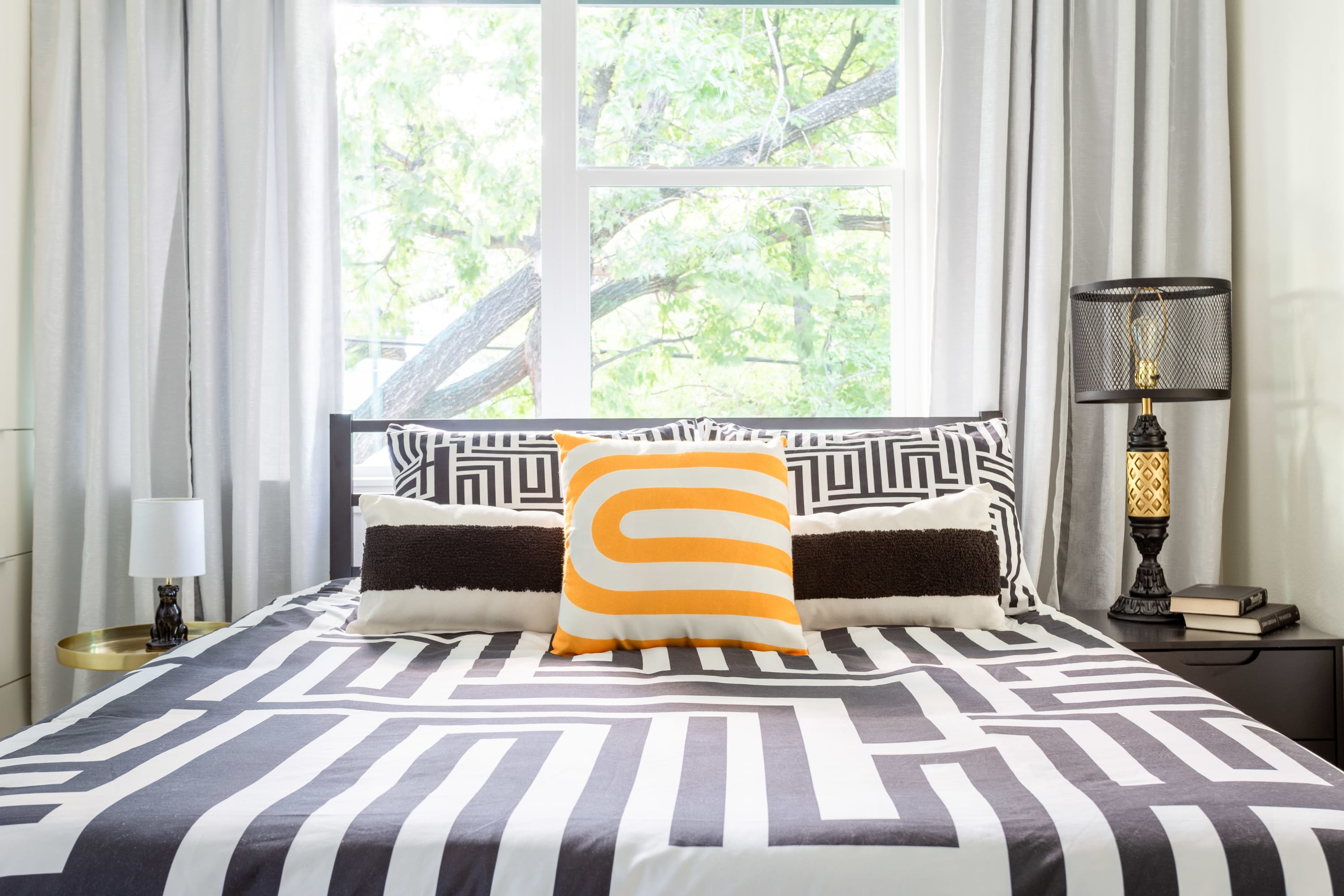 Close-up view of a modern bedroom bed with black and white geometric bedding and a vibrant orange accent pillow, positioned against a large window with white curtains overlooking lush trees. Two black and gold table lamps flank the bed on dark nightstands.