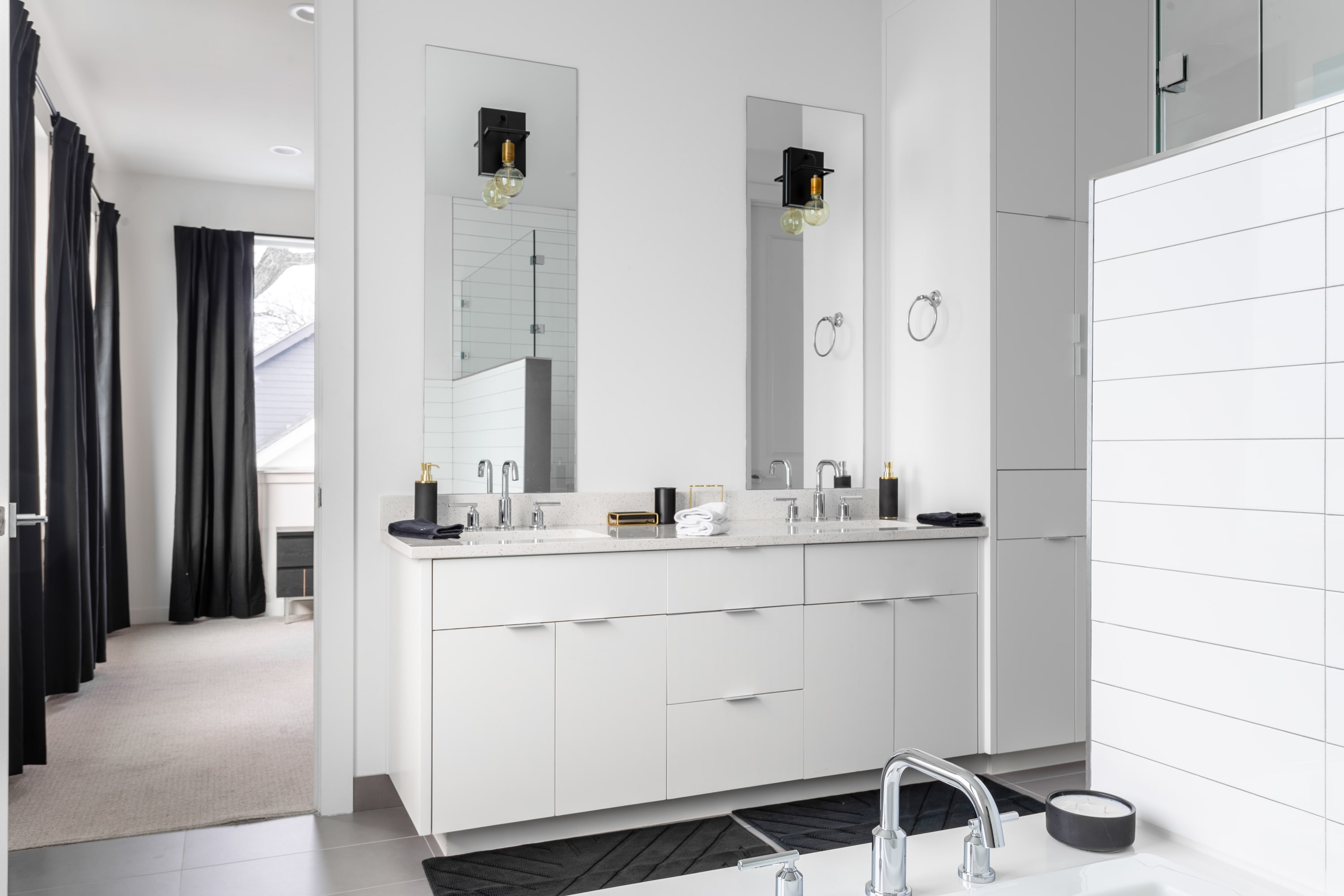 Modern bathroom featuring a white double vanity with two rectangular mirrors, black sconces, and chrome fixtures, leading into a bedroom with dark curtains.