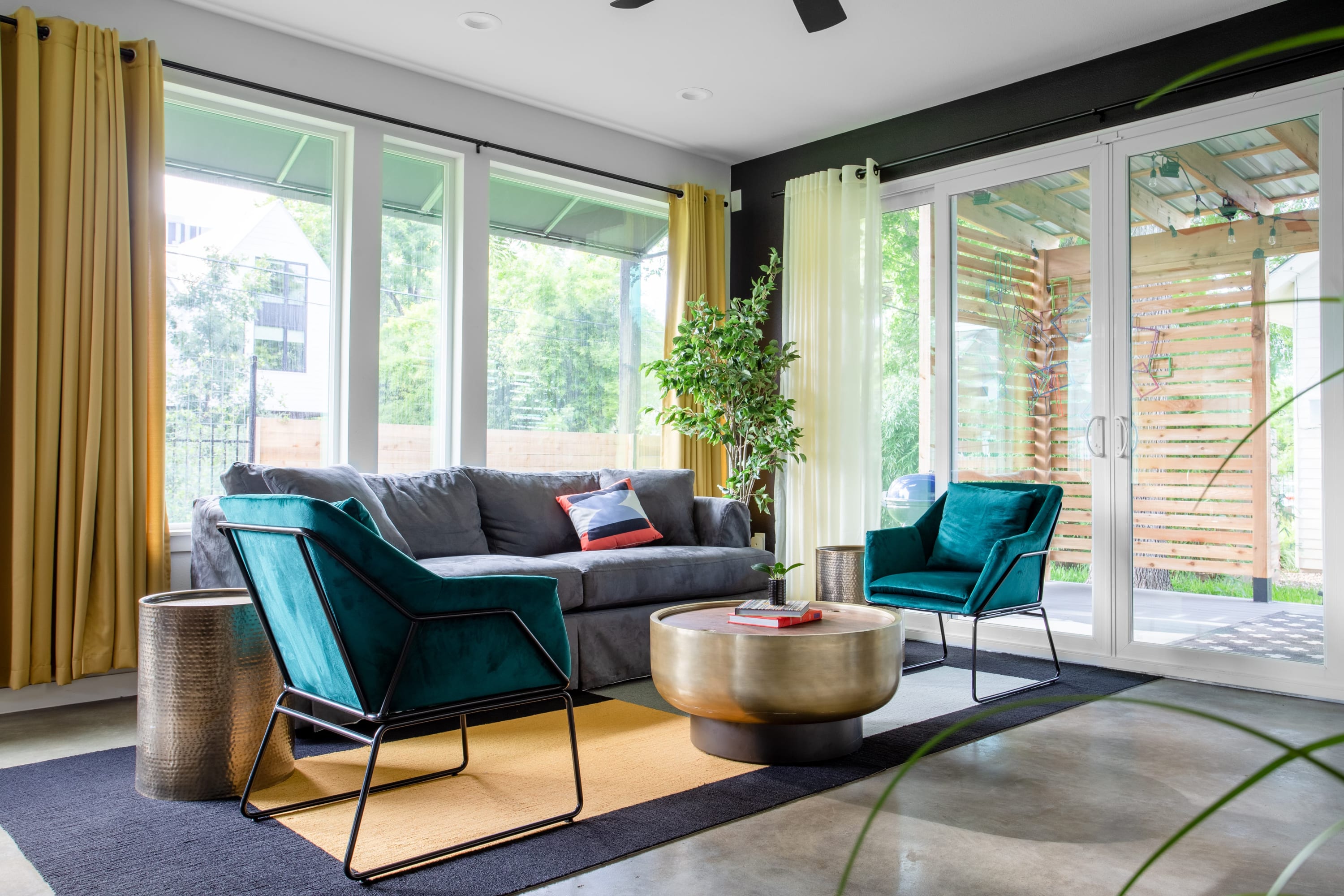 A bright living room with large windows and sliding glass doors features a gray sofa, two teal velvet armchairs, and a round coffee table on a colorful rug, leading to an outdoor patio.
