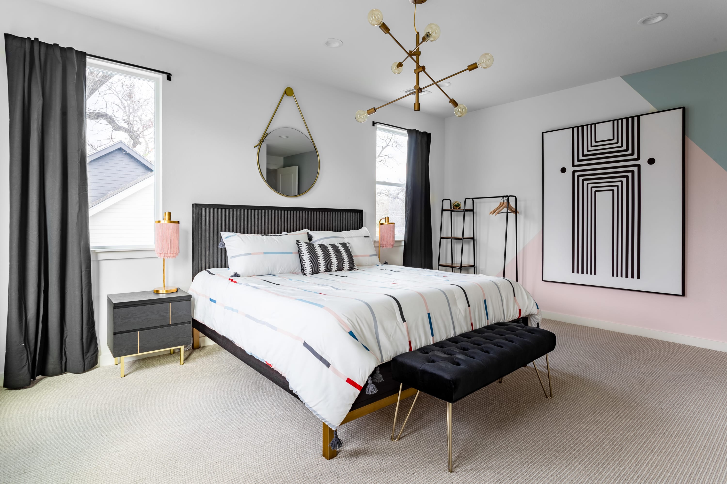 Stylish bedroom with a king-sized bed, black headboard, and a large abstract artwork on a feature wall with geometric pink and green color blocks. A modern chandelier and black curtains complete the contemporary look.