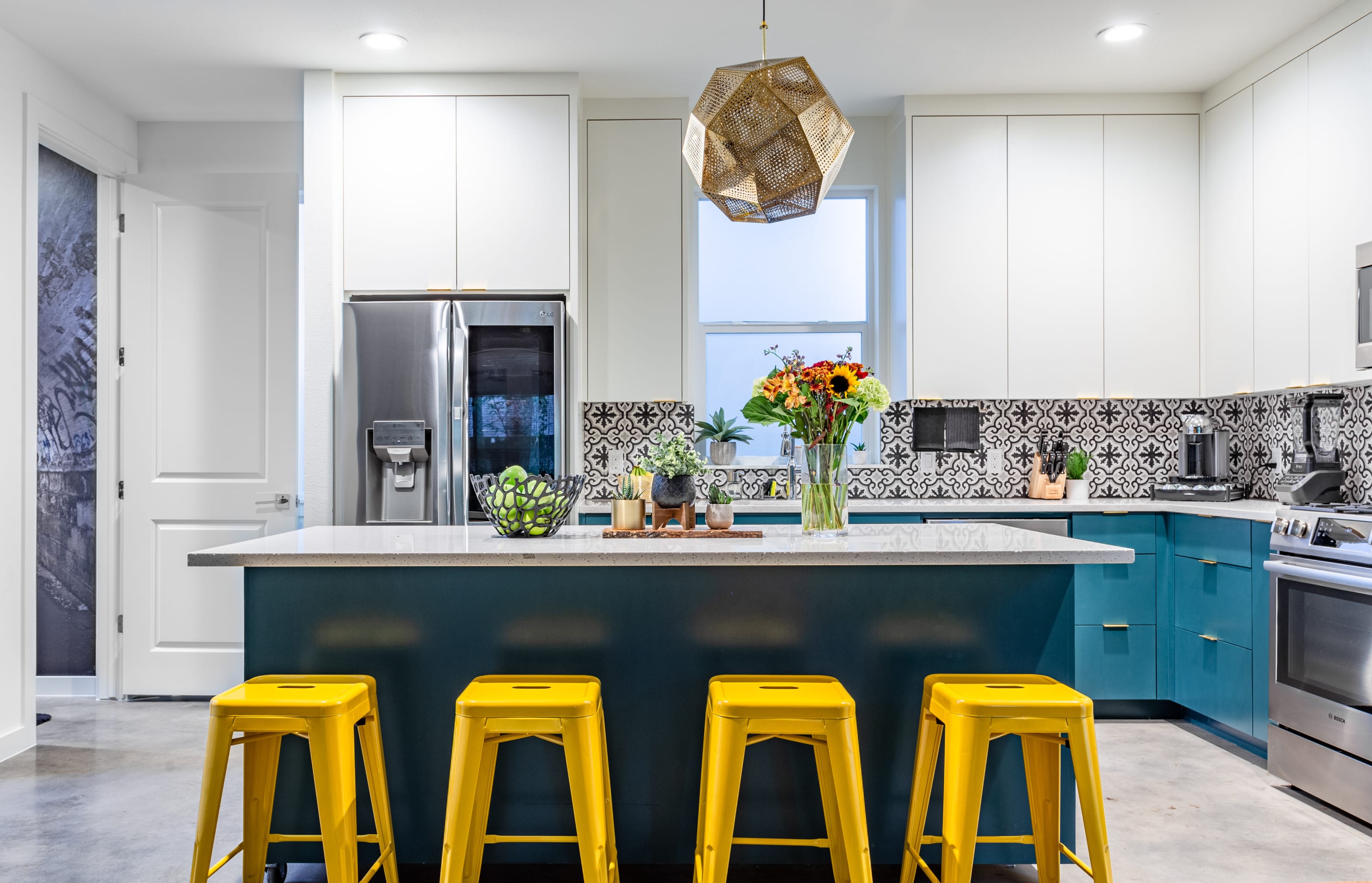Stylish kitchen featuring a large island with bright yellow bar stools, white quartz countertops, and teal lower cabinets. A unique geometric pendant light hangs above, and a vase of fresh flowers adds a pop of color.