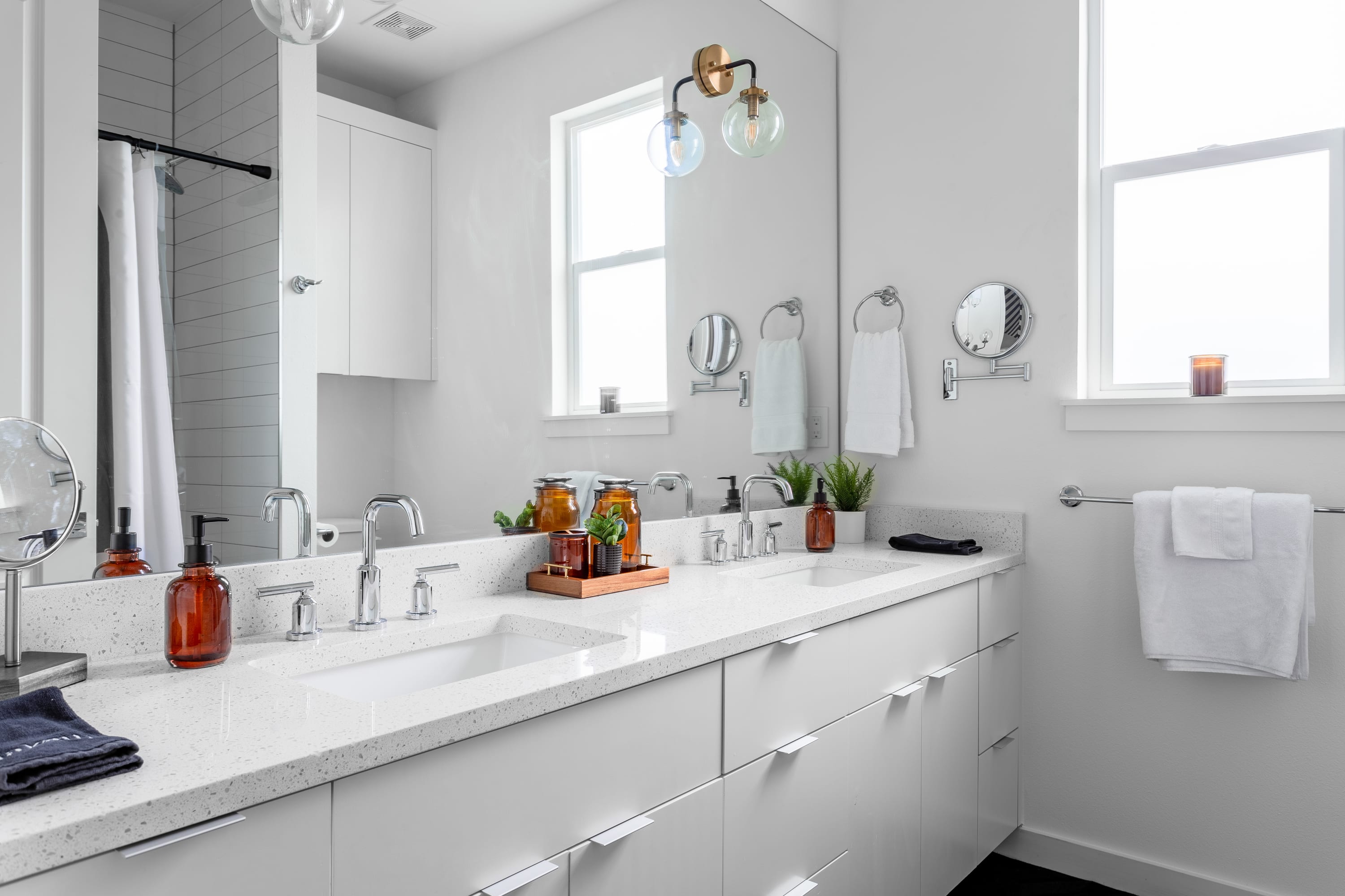 A spacious modern bathroom features a long white double vanity with two sinks, large mirrors, and chrome fixtures. Two windows provide natural light, and the room is decorated with small green plants and amber-colored soap dispensers.
