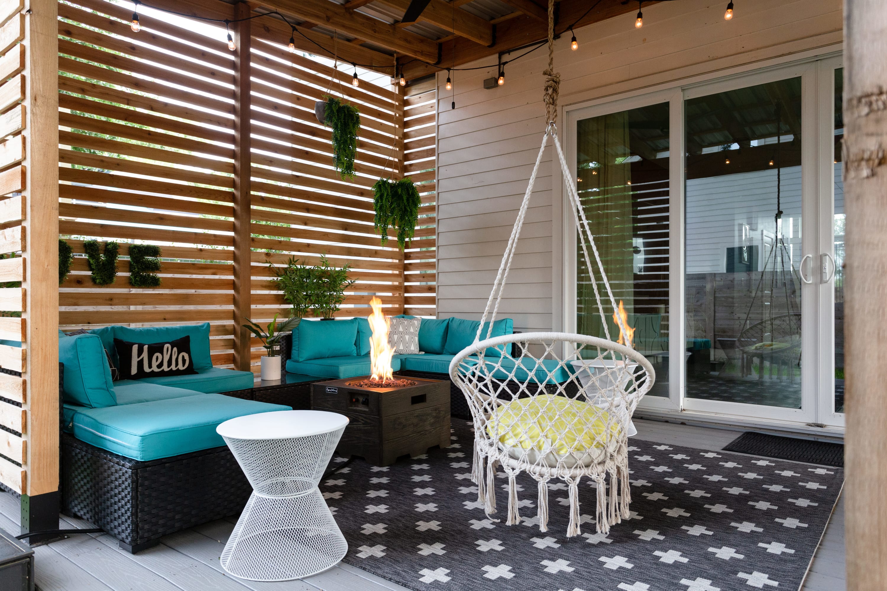 A stylish covered patio features teal outdoor sofas and a fire pit, with a white macrame swing chair and a geometric rug, offering a cozy outdoor living space.