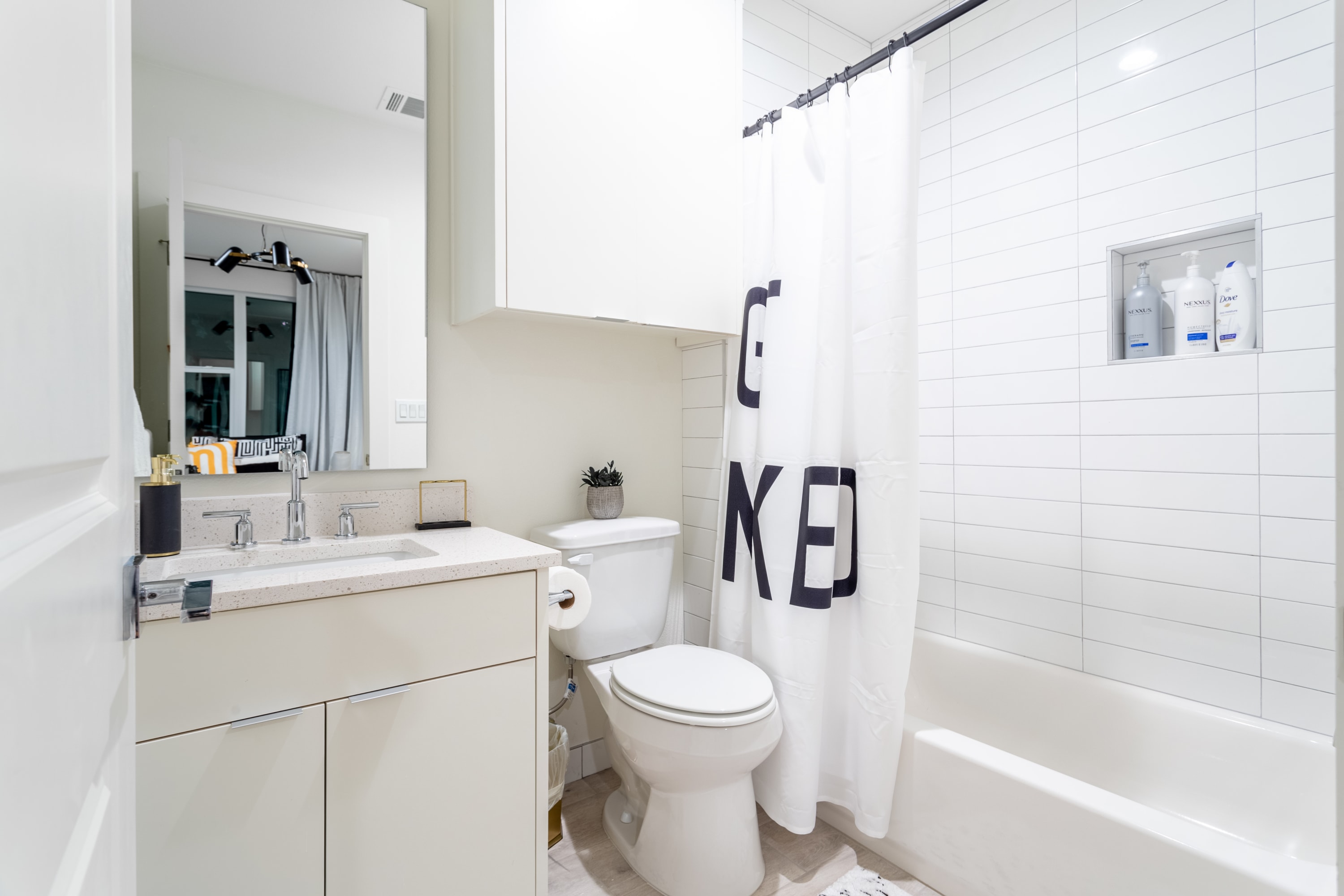 Bright, modern bathroom featuring a white subway-tiled shower with a white shower curtain displaying bold black text, a white vanity with a stone countertop and chrome faucet, and a toilet. A recessed shelf in the shower holds toiletries.