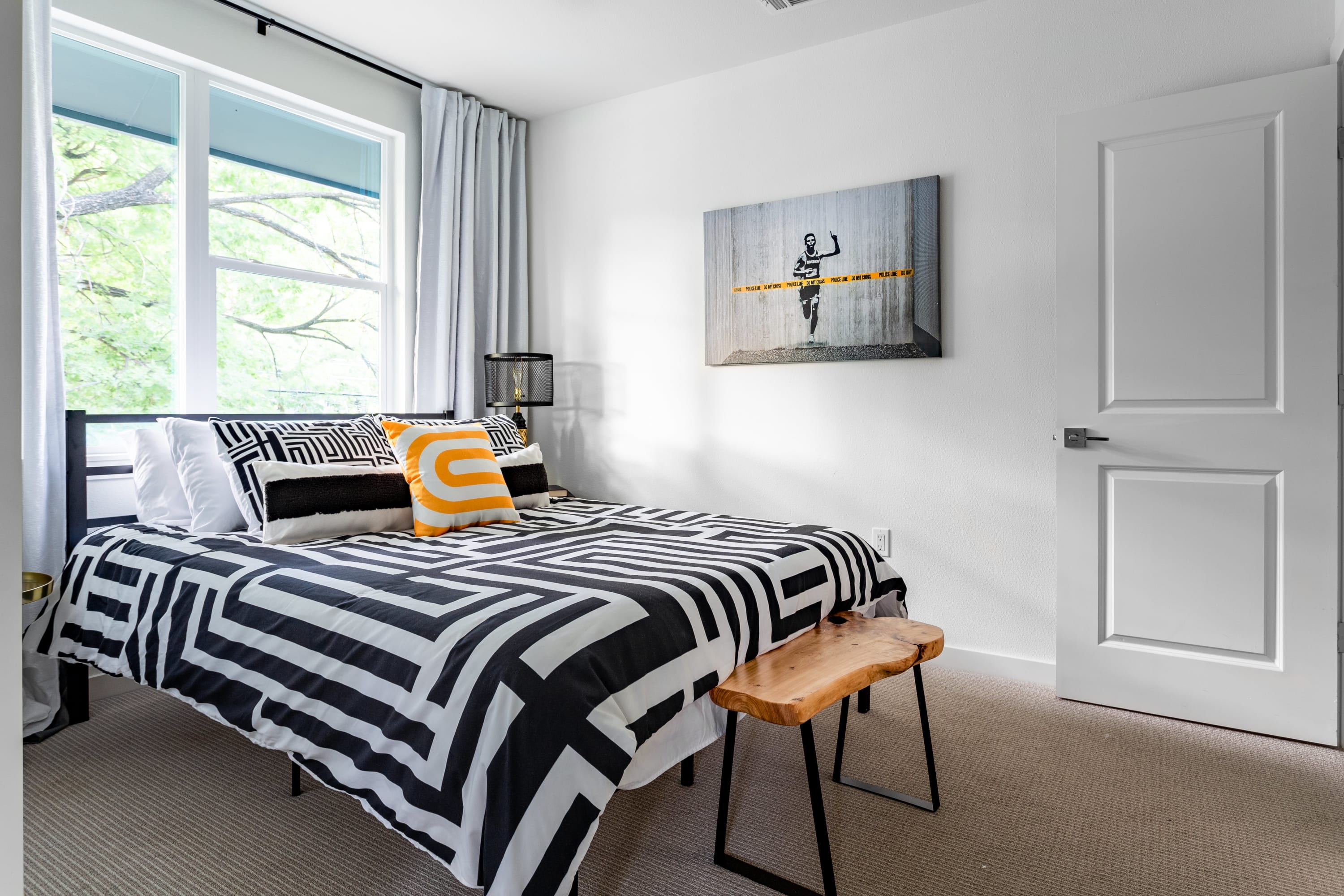 Modern bedroom featuring a bed with striking black and white geometric bedding, an orange accent pillow, and a large window offering a view of trees. A contemporary art piece hangs on the white wall, complemented by a small wooden bench.