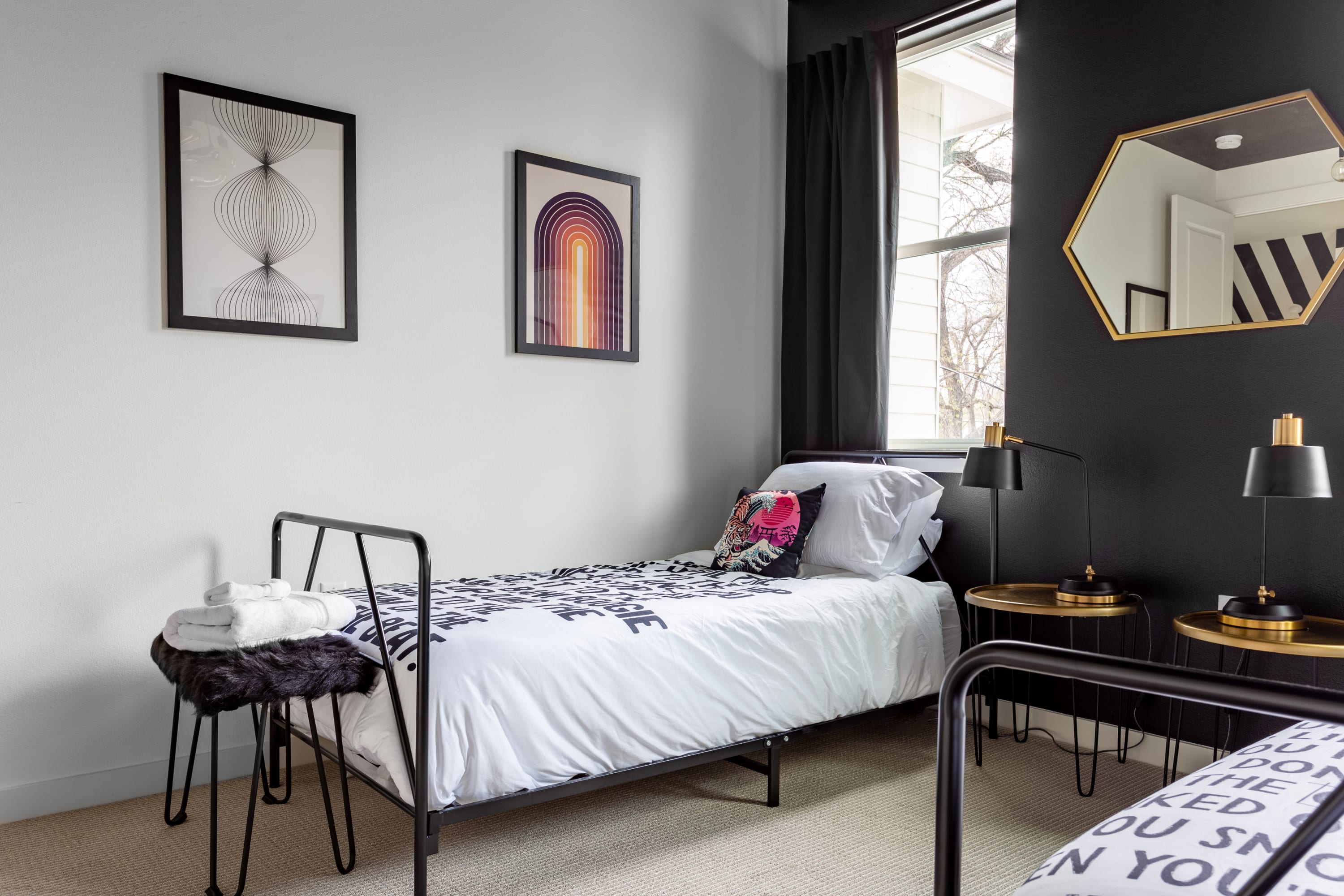A vibrant bedroom showcases two black metal-framed twin beds with white bedding featuring black text patterns and a colorful abstract pillow. The room has a black accent wall, white walls with framed abstract art, and a gold hexagonal mirror reflecting the space.