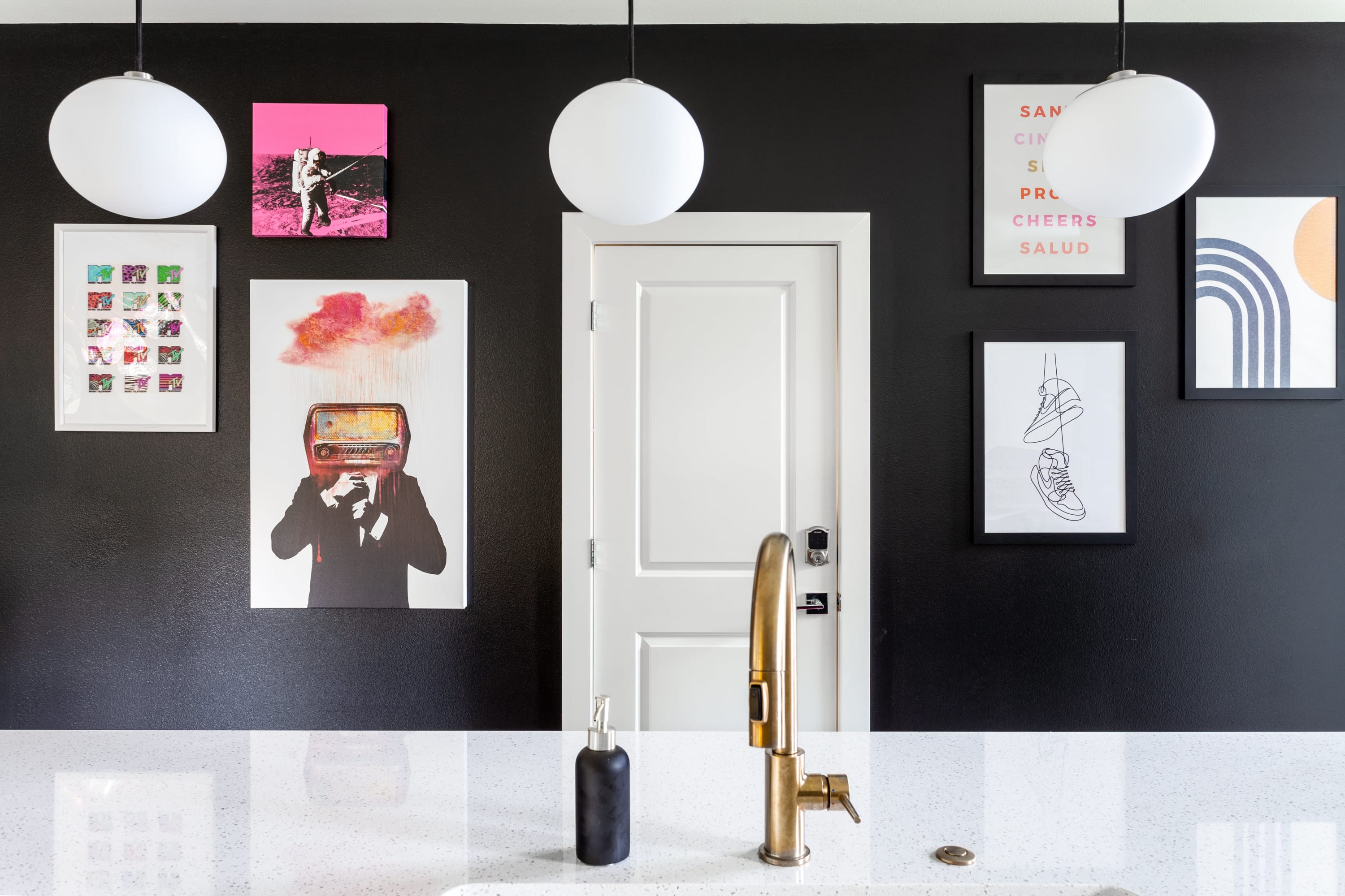 Close-up of a kitchen sink area with a modern gold faucet and white speckled quartz countertop, set against a dramatic black wall adorned with eclectic framed art and a white door.