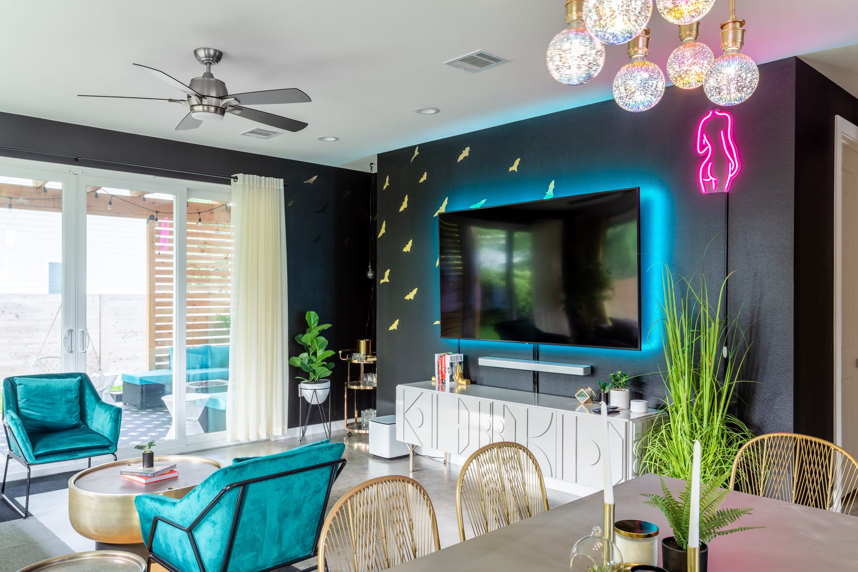A contemporary living space features a large wall-mounted TV with blue neon backlighting, a white media console, and teal velvet chairs, with a unique bubble chandelier overhead.