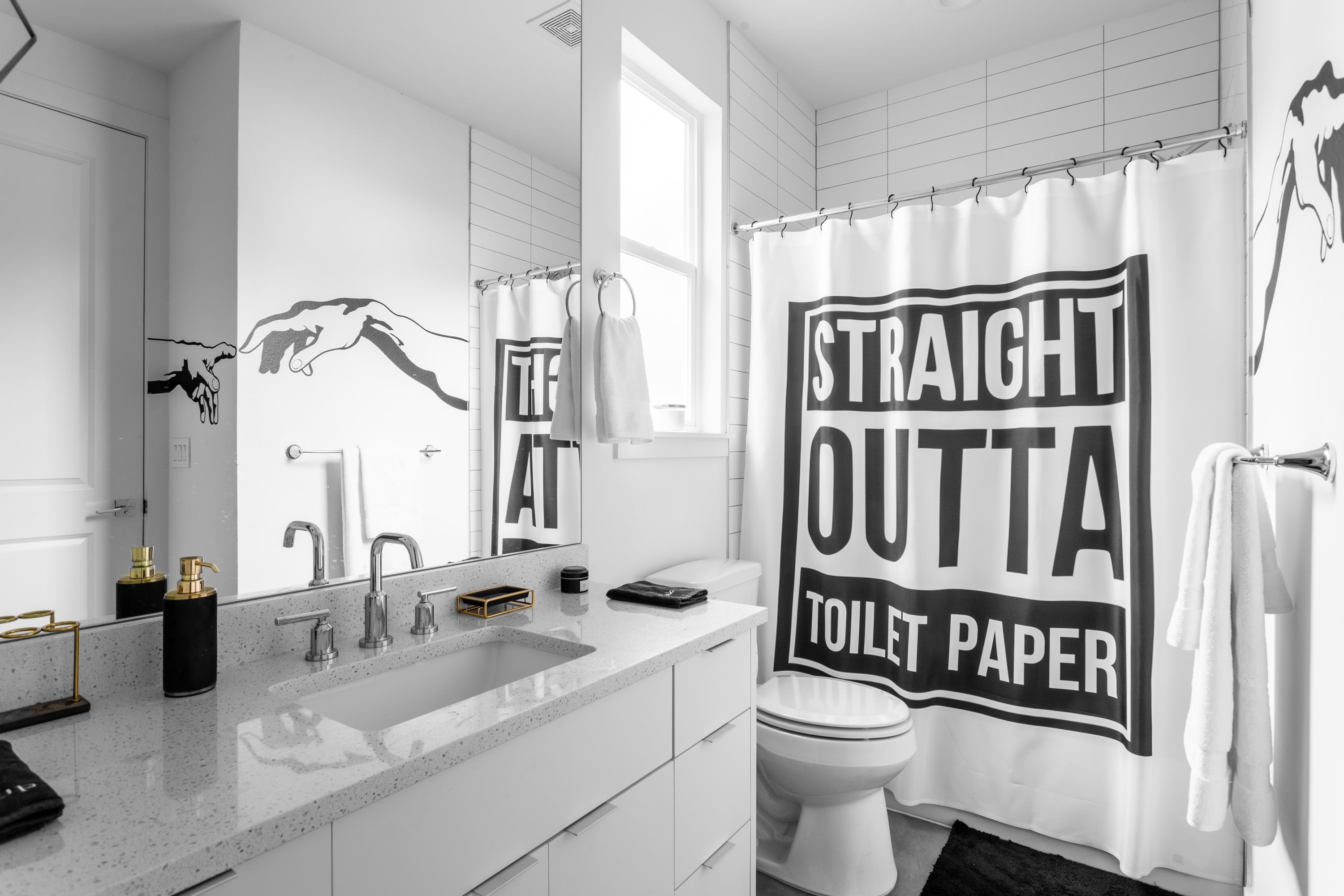 A modern white bathroom features a large vanity with a single sink, a wide mirror, and black and white wall art depicting hands. A toilet is visible next to a shower with a "STRAIGHT OUTTA TOILET PAPER" shower curtain.