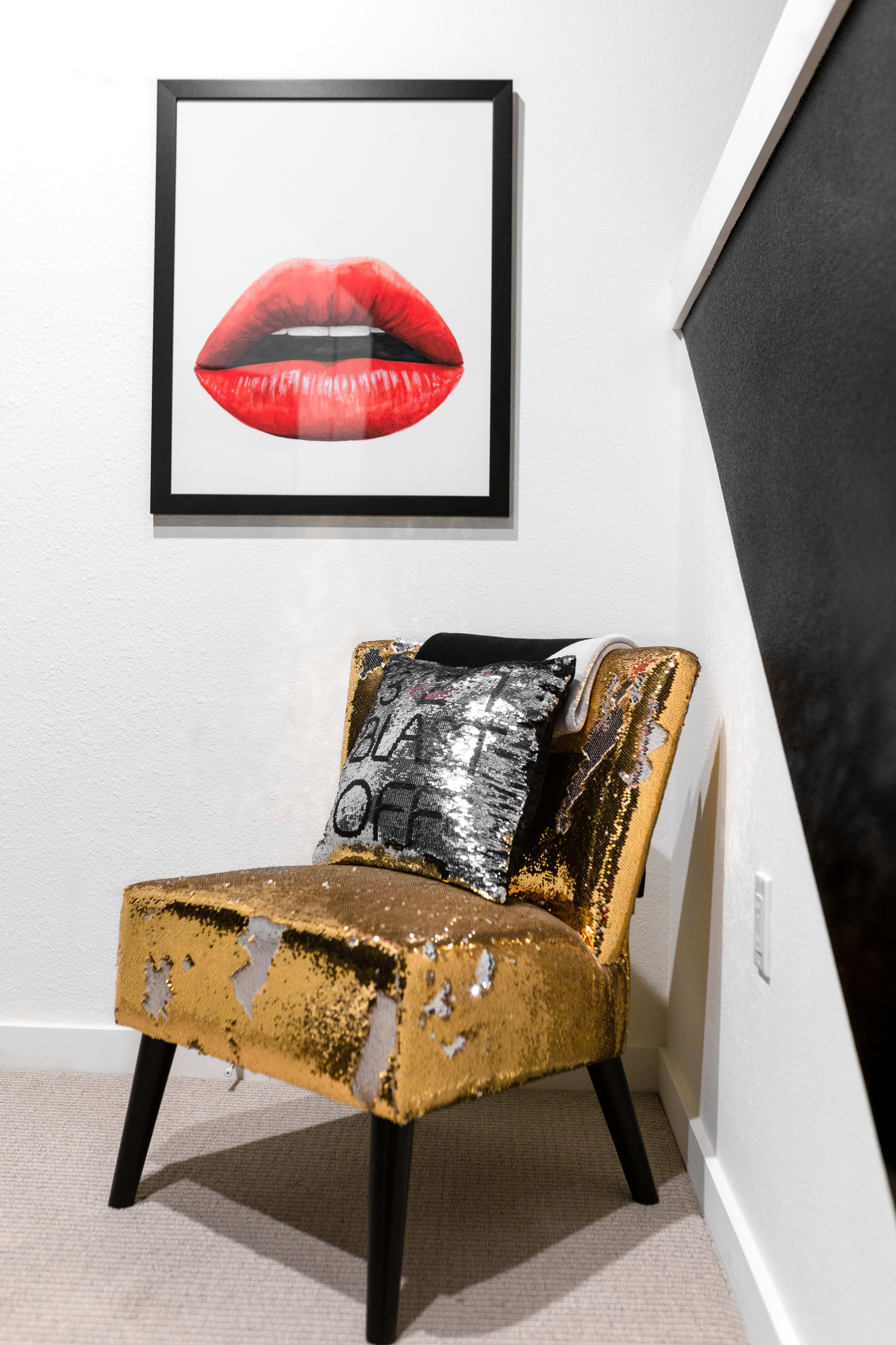 Close-up of a gold sequined chair with a silver sequined pillow, positioned beneath a framed pop art print of red lips, against a white wall.