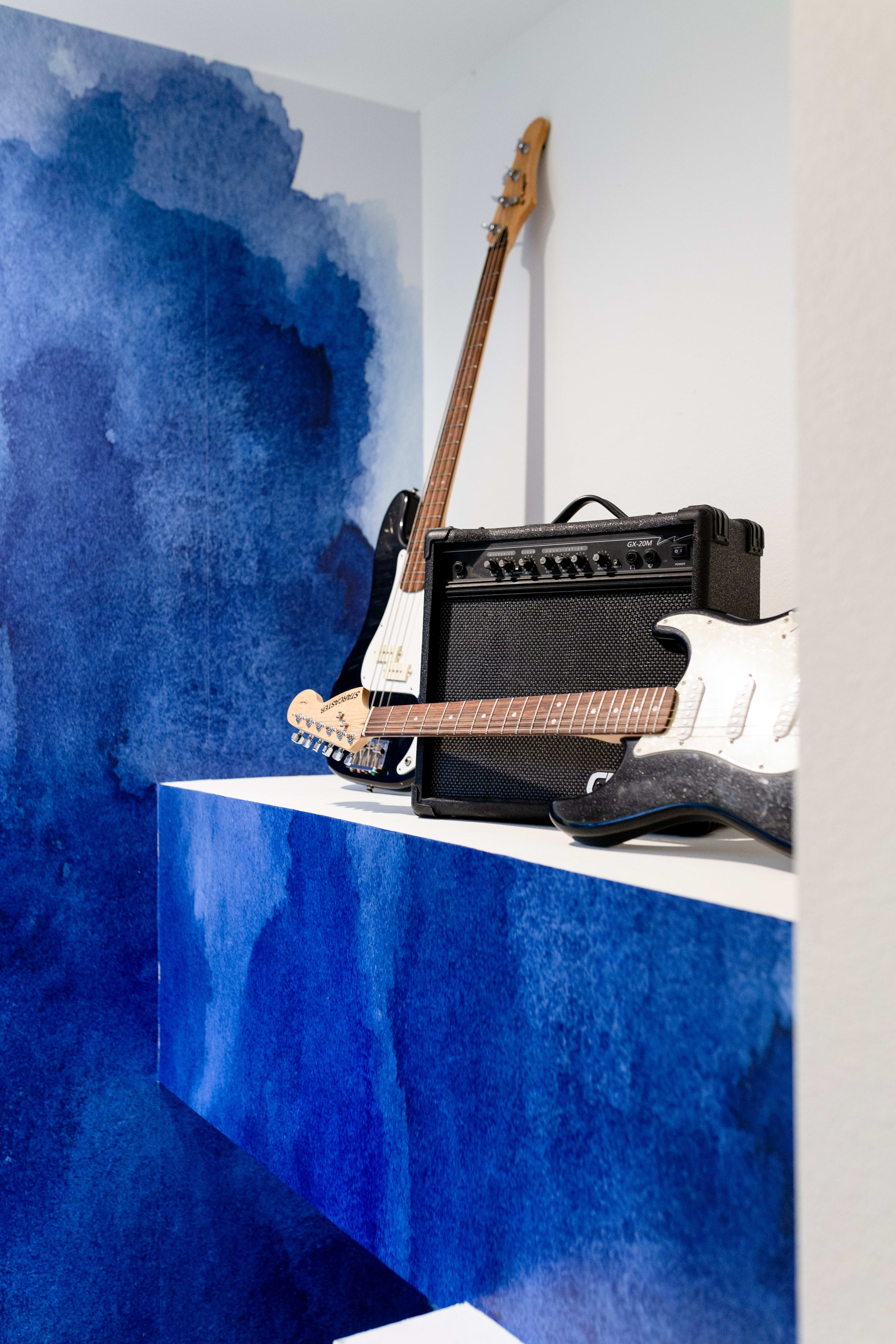 A vibrant music corner features a blue watercolor-patterned wall and a white platform, holding two electric guitars (one bass, one standard) and a black guitar amplifier.