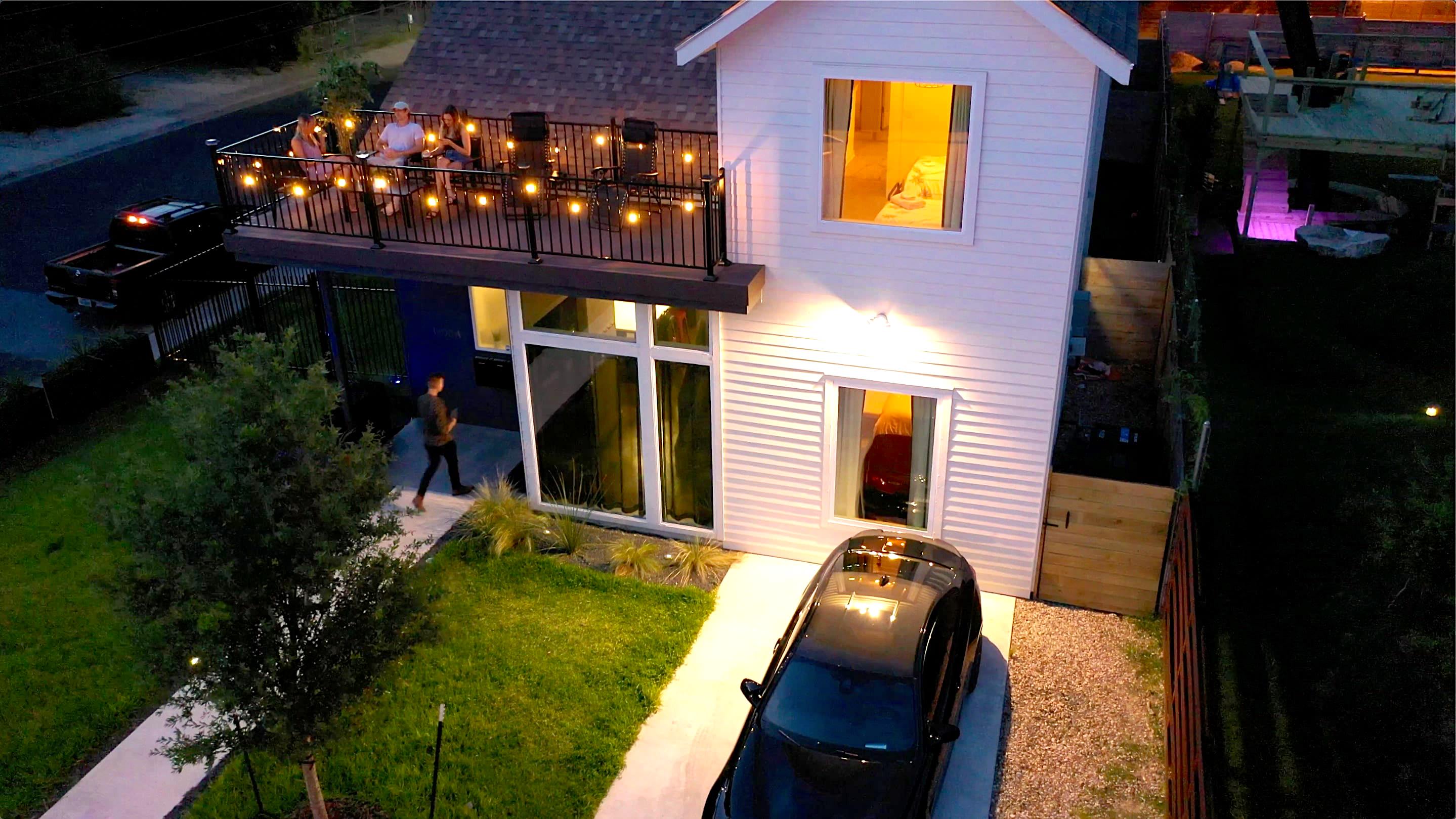 Aerial view of a modern white house at dusk, featuring a second-story balcony with string lights and people gathered. A car is parked in the driveway, and a small lawn is visible in front.