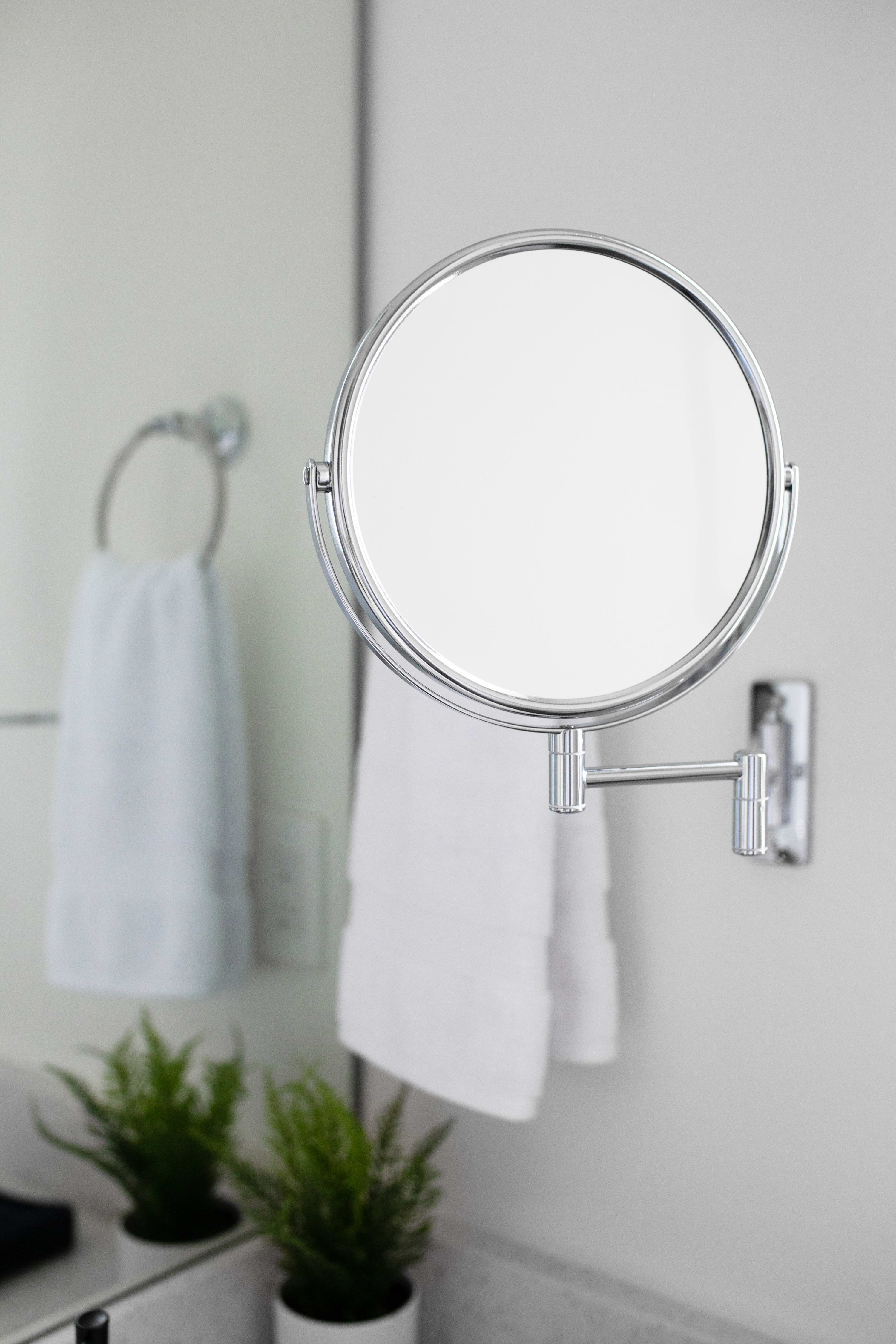 A close-up of a round, extendable magnifying mirror mounted on a white wall in a bathroom, with a white hand towel hanging nearby and green potted plants reflected in the background.
