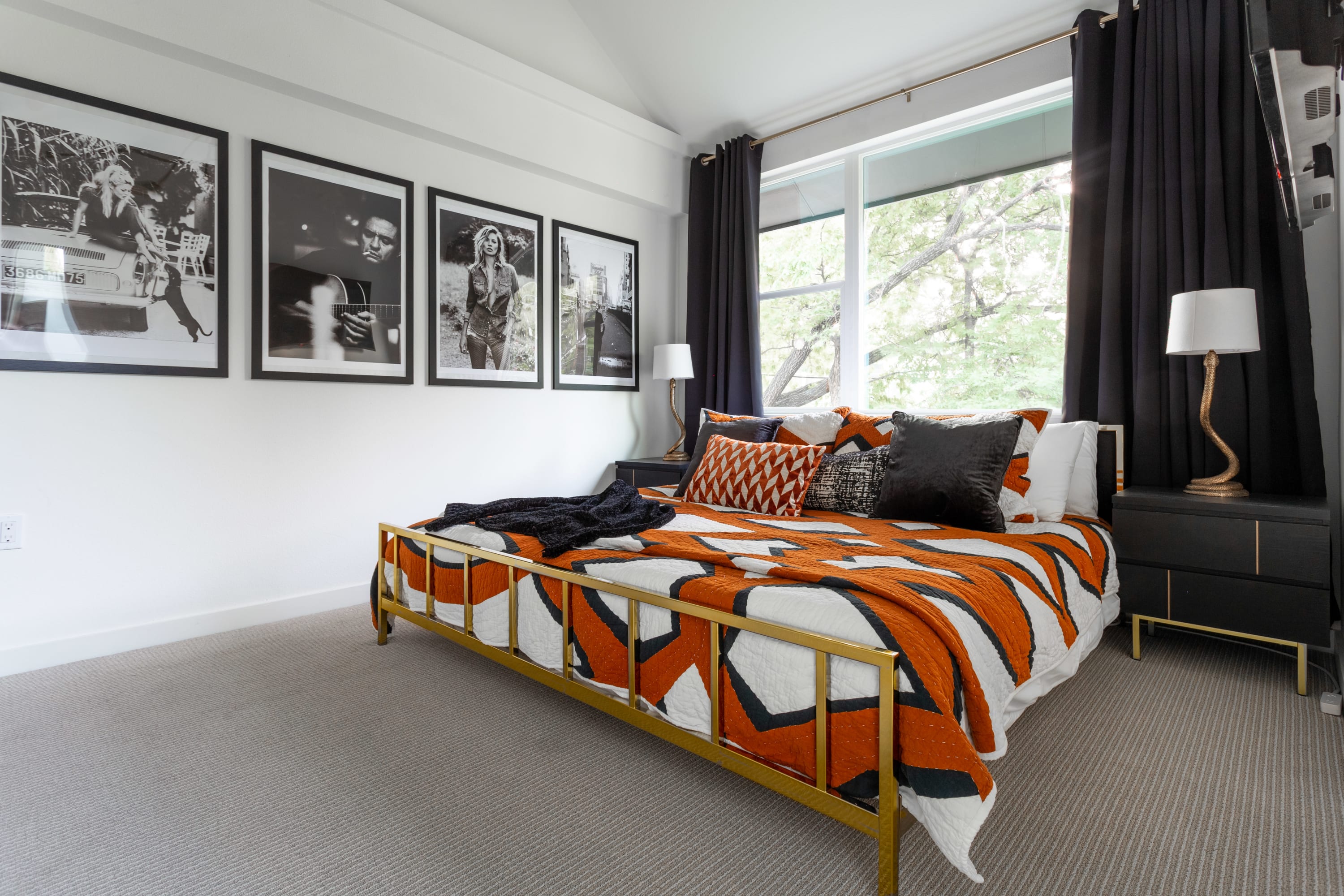 Spacious bedroom featuring a gold metal bed frame with an orange and black patterned quilt, flanked by black nightstands, and a gallery wall of black and white framed art.