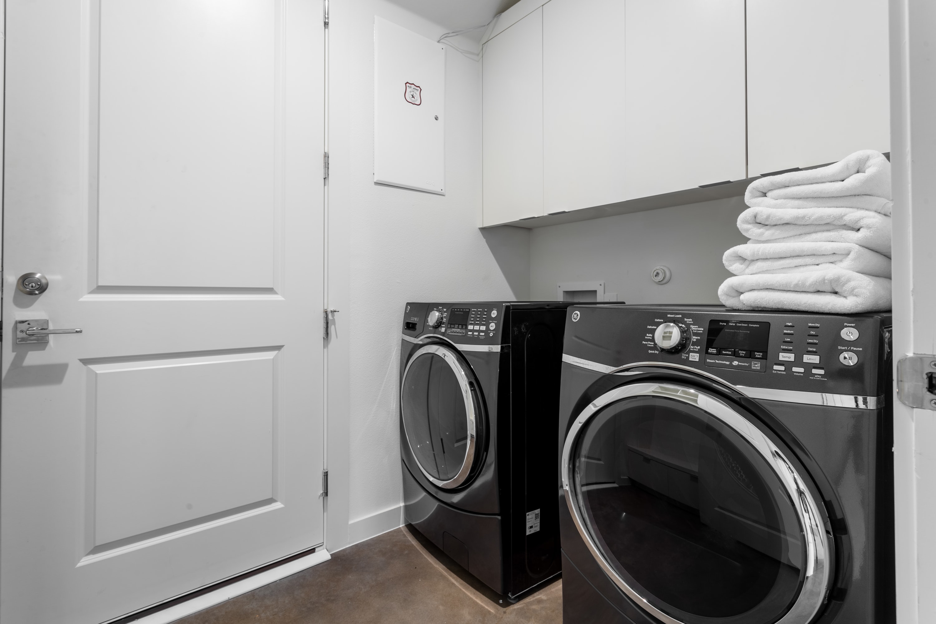 Functional laundry room equipped with two sleek black front-loading washing machines, stacked white towels, and ample white cabinetry for storage. A white door and light-colored walls complete the clean and practical space.