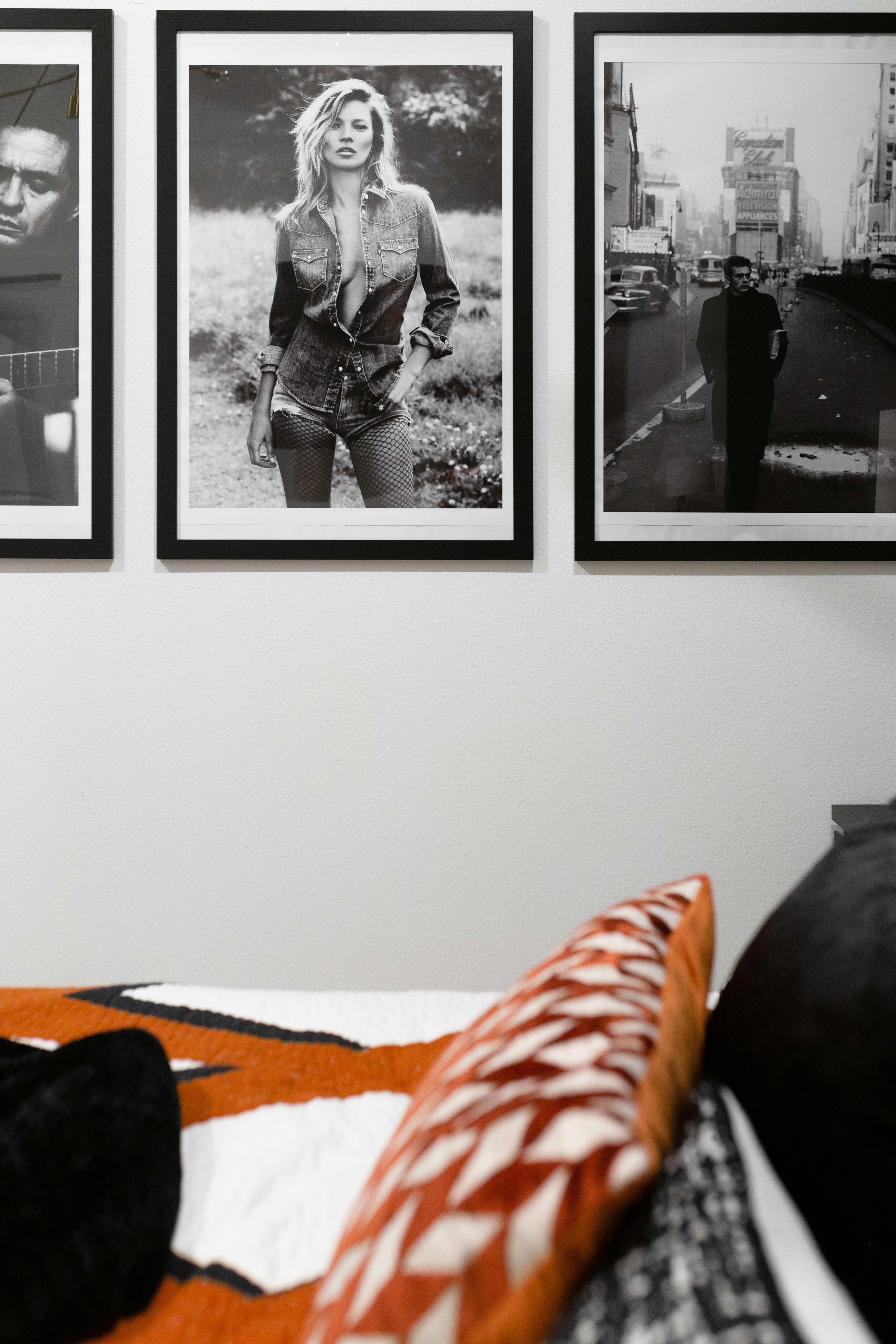 Close-up of a gallery wall in a bedroom, showcasing three black and white framed photographs, including one of Kate Moss, above a bed with an orange and black patterned quilt.