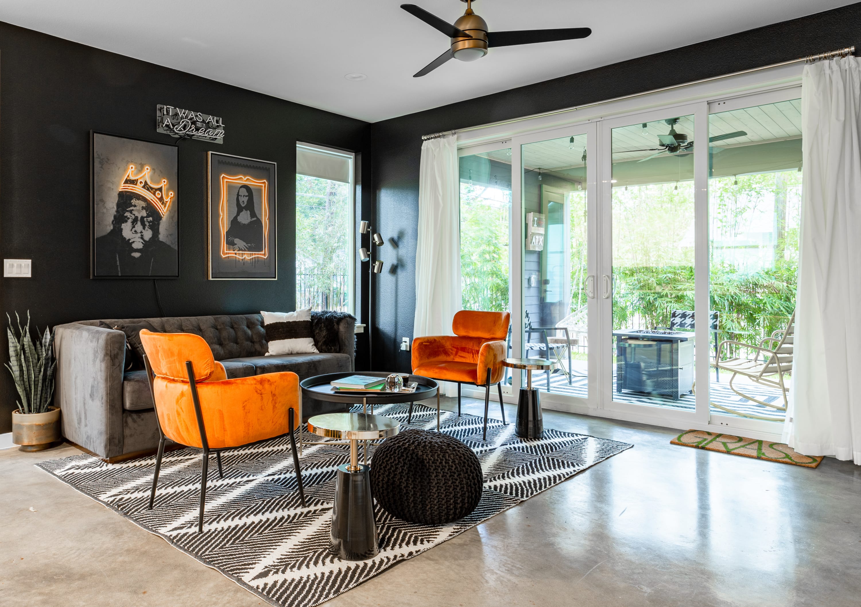 A modern living room with dark walls and concrete floors features a gray sofa, two orange velvet armchairs, and a round coffee table on a black and white rug, with sliding doors to a patio.