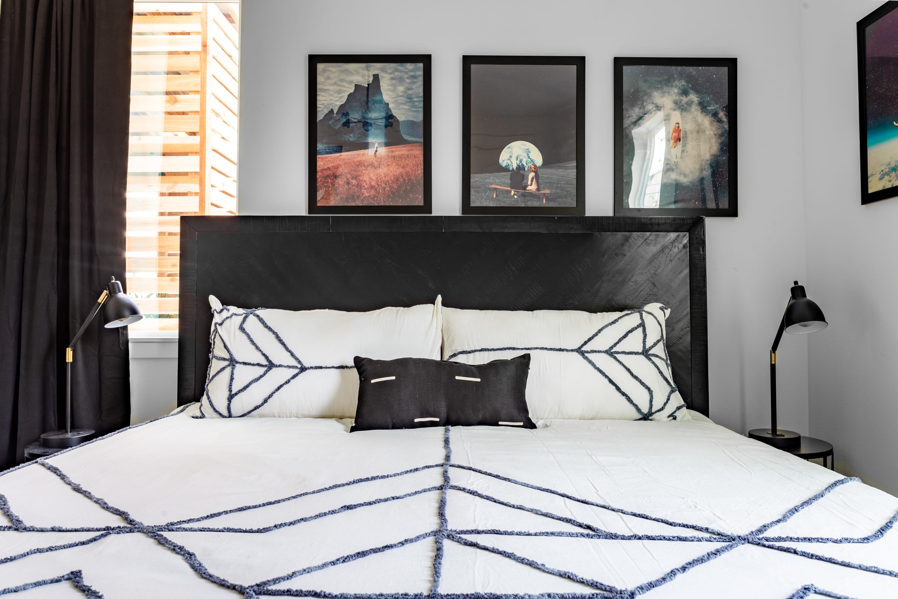 Close-up of a king-sized bed in a modern bedroom, featuring white bedding with intricate black geometric embroidery and a dark rectangular accent pillow. A dark wooden headboard frames the bed, flanked by black table lamps on matching nightstands.