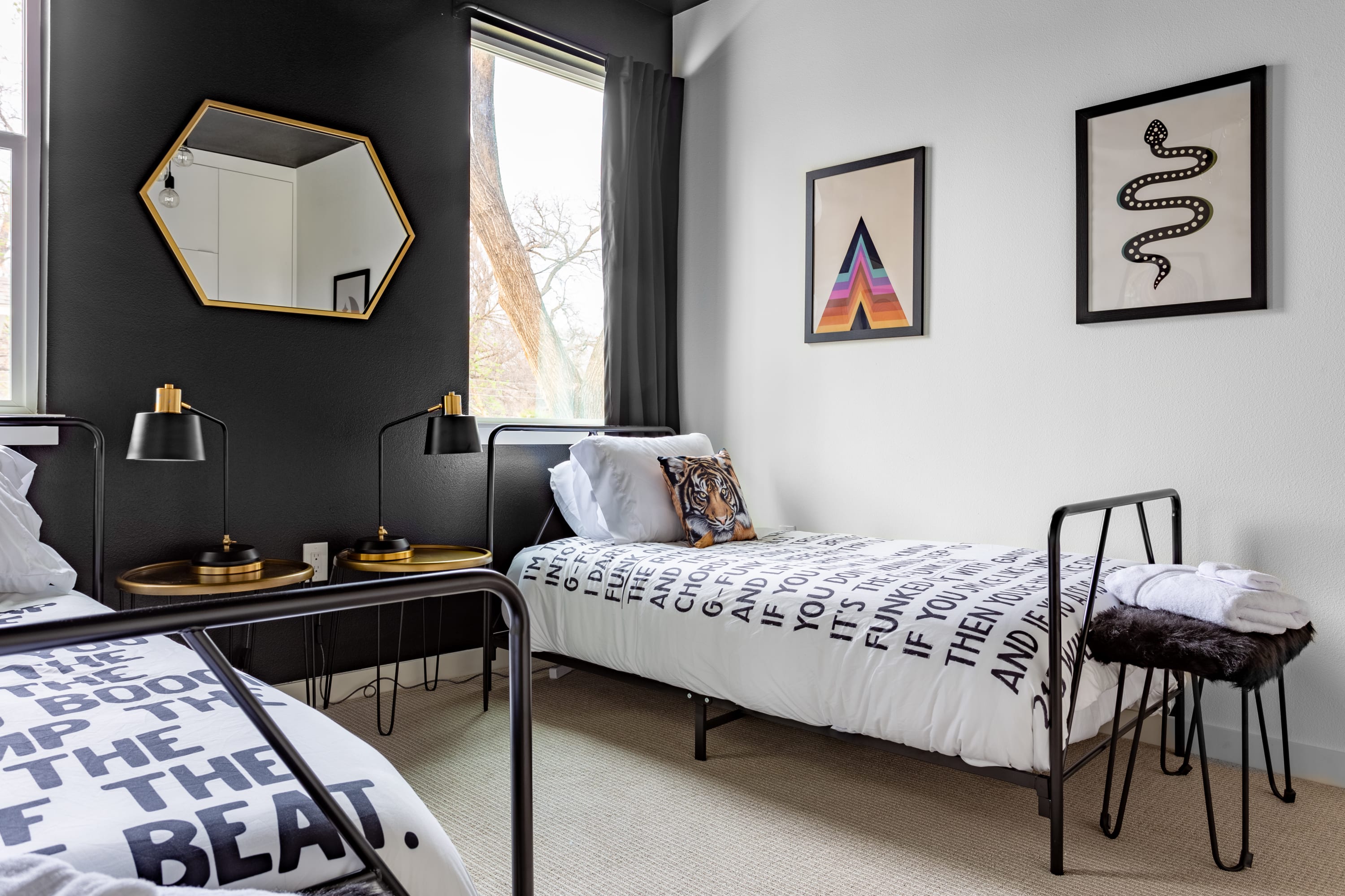 A stylish bedroom with two black metal-framed twin beds featuring white bedding with bold black text patterns and a tiger print pillow. A black accent wall contrasts with white walls, adorned with colorful geometric and snake art prints, and a gold hexagonal mirror.