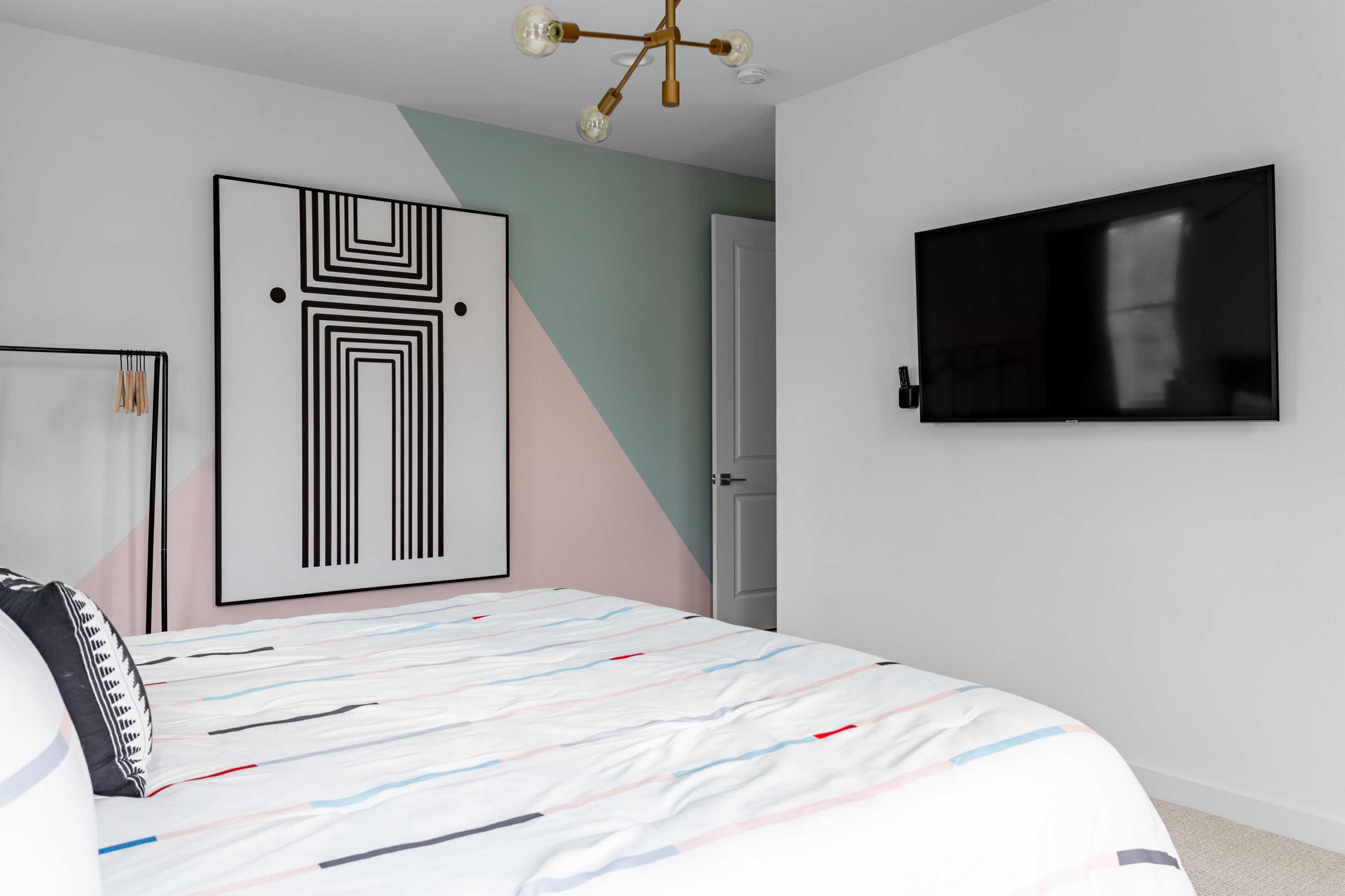 Detail shot of a bedroom's feature wall, showcasing a large black and white abstract artwork against a backdrop of geometric pink and mint green color blocks. A wall-mounted television is visible on the adjacent white wall.