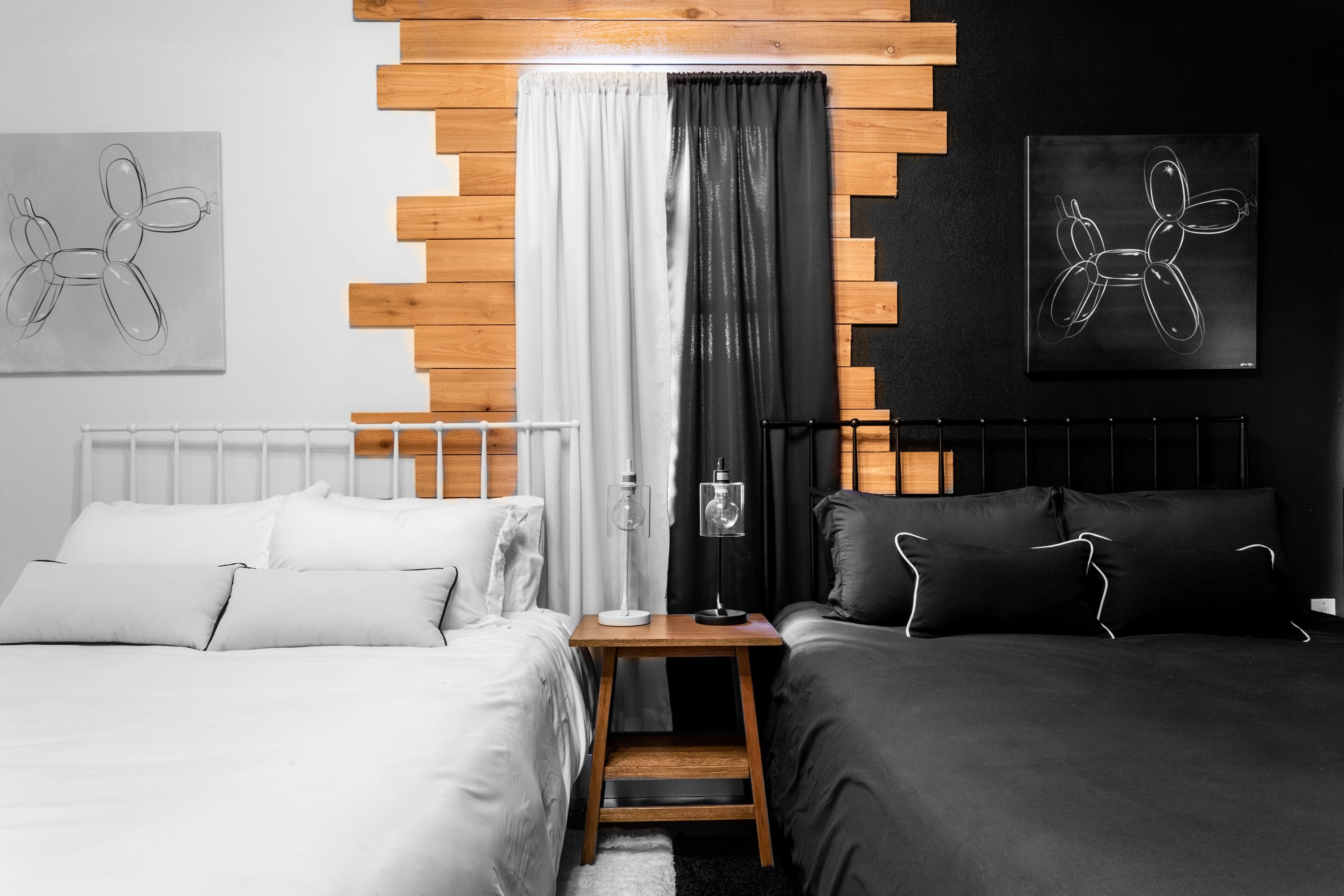 Striking bedroom with a split black and white design, featuring two distinct beds and a central wooden accent wall. Each side is adorned with a "balloon dog" artwork, creating a playful yet modern aesthetic.