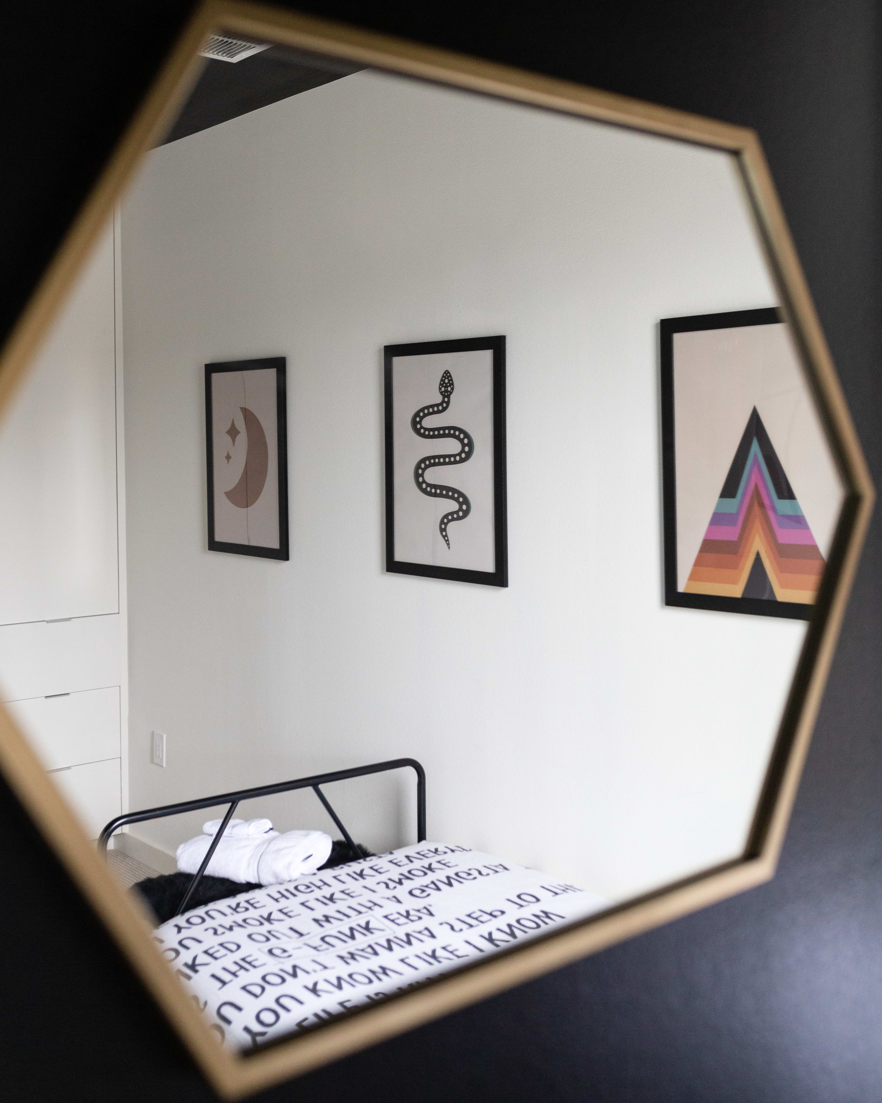 A close-up view through a gold hexagonal mirror, reflecting a portion of the bedroom with white walls, framed art prints including a moon, a snake, and colorful geometric shapes, and a black metal-framed twin bed with patterned bedding.