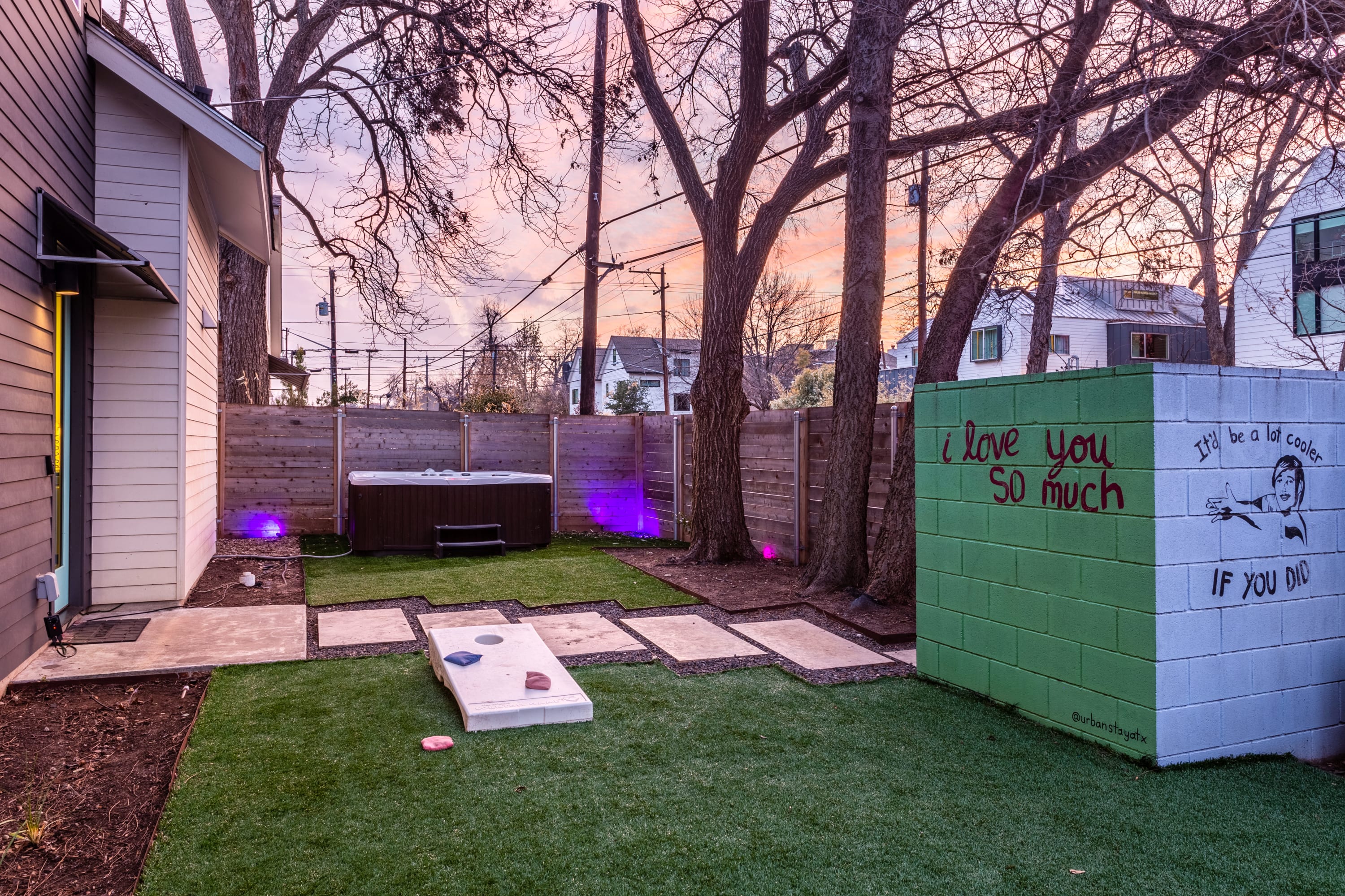 Fenced backyard with artificial turf, a hot tub, and a colorful block wall mural featuring "I love you so much" and "It'll be a lot cooler if you did" text. Purple uplights illuminate the fence and hot tub area.