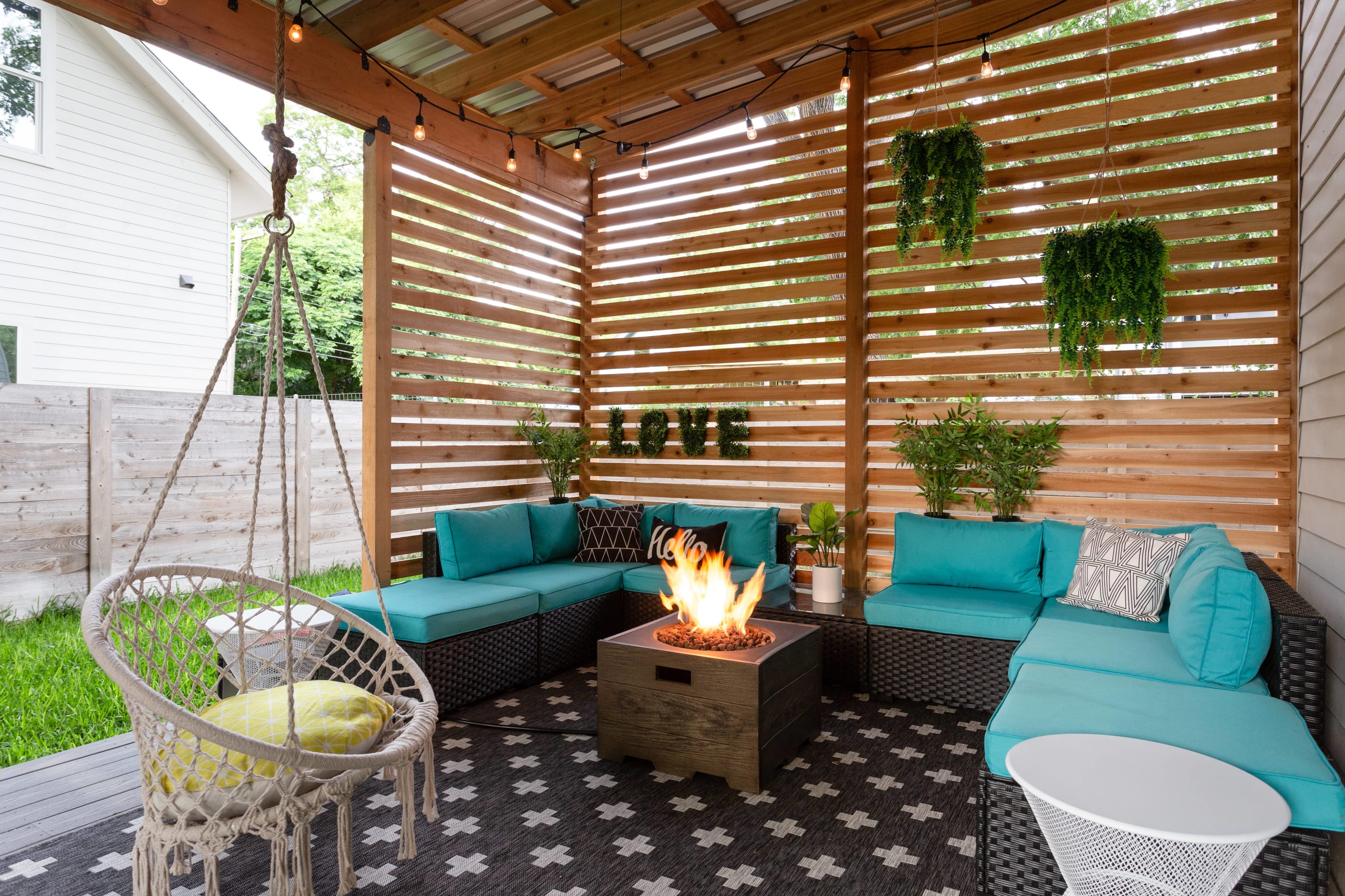 Covered outdoor patio with a slatted wooden roof and walls, featuring a turquoise sectional sofa arranged around a square fire pit. String lights illuminate the space, and hanging plants add a touch of greenery.