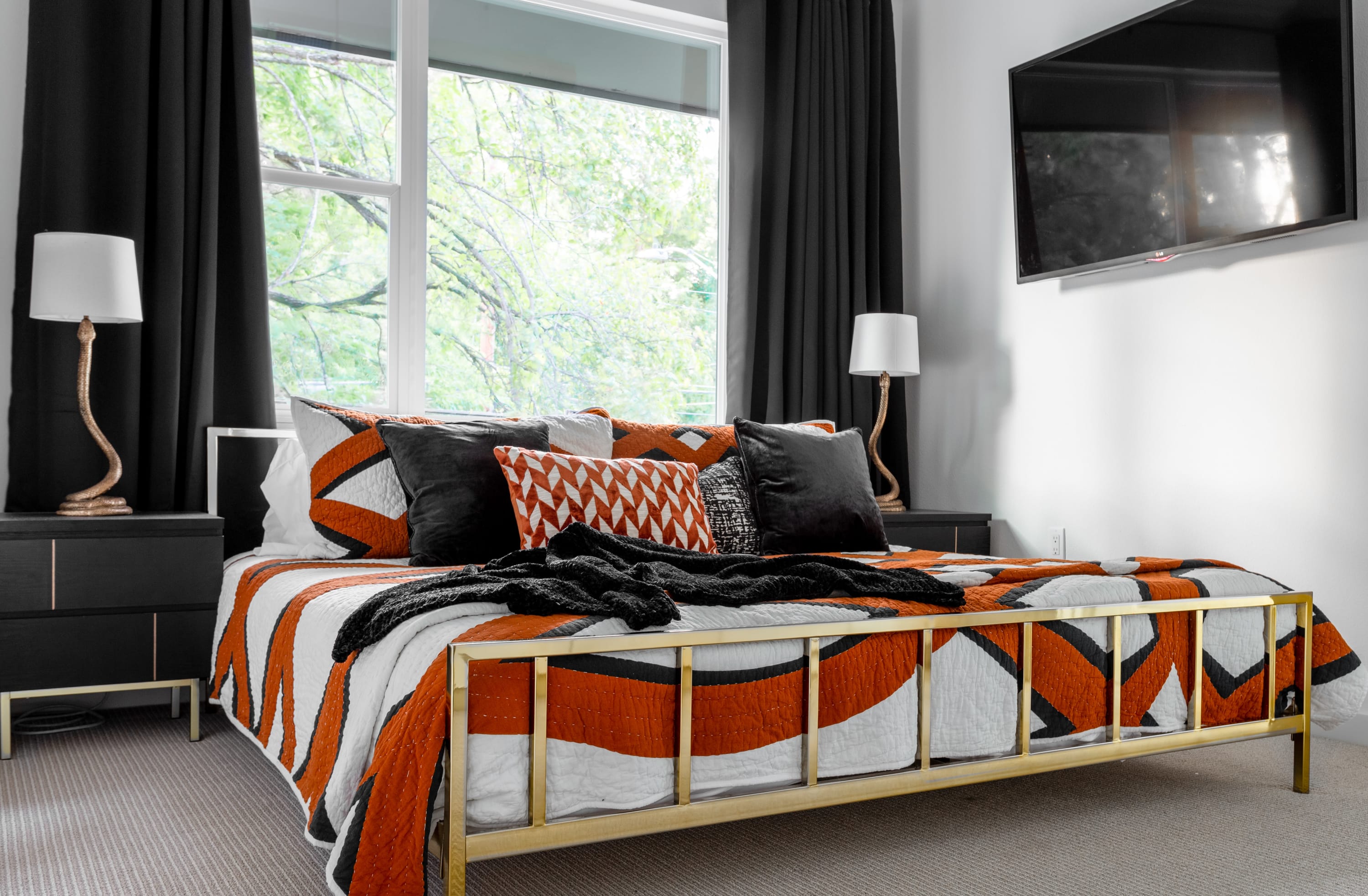 Vibrant bedroom with a gold metal bed frame, an orange, white, and black patterned quilt, and large windows with black curtains overlooking trees.