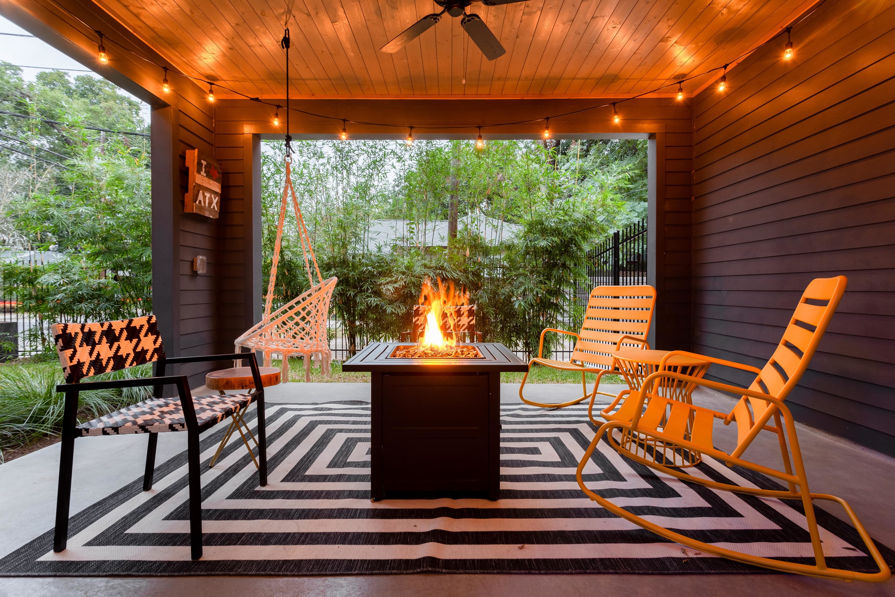 A covered outdoor patio showcases a central fire pit with flames, surrounded by yellow metal rocking chairs and a black and white striped rug, under a wooden ceiling with string lights.