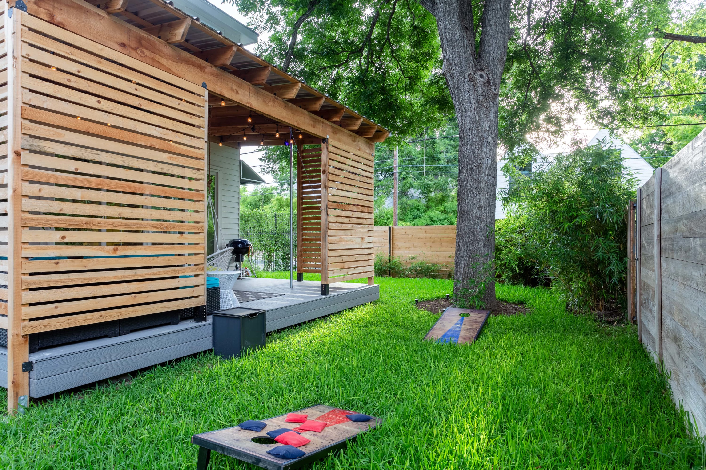 Lush green backyard featuring a wooden slatted covered patio with a BBQ grill and a cornhole game set up on the grass. Mature trees provide shade, and a wooden fence encloses the area.