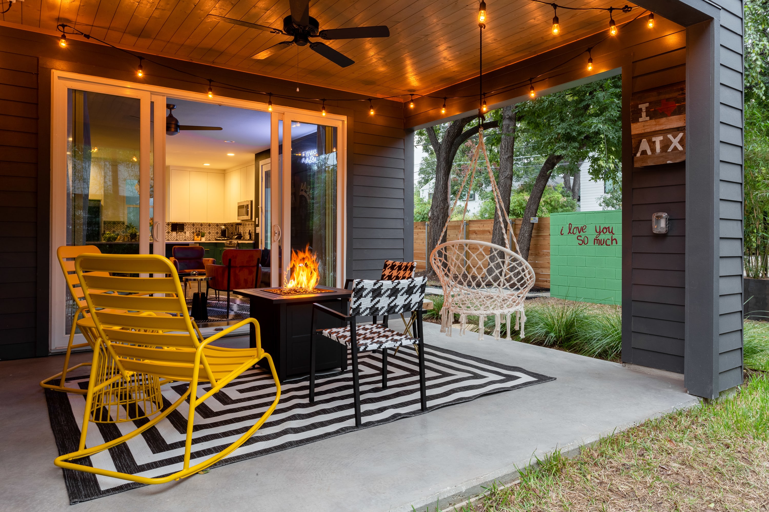 A modern covered patio features a central fire pit, yellow metal rocking chairs, a black and white striped rug, and string lights overhead, with a view into an indoor living space.