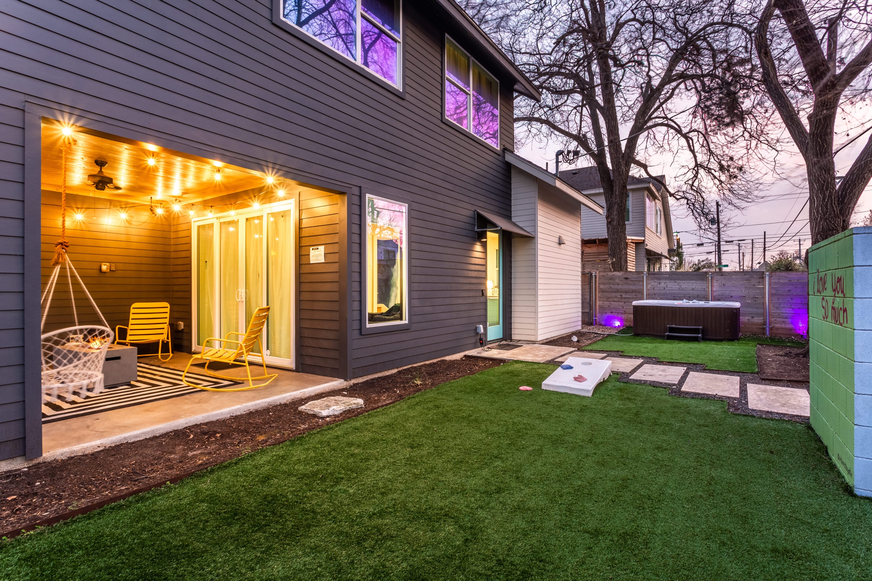 Rear view of a dark gray house with a covered patio featuring string lights, a swing chair, and yellow rocking chairs. The backyard has artificial turf, stepping stones, a cornhole game, and a hot tub in the distance.