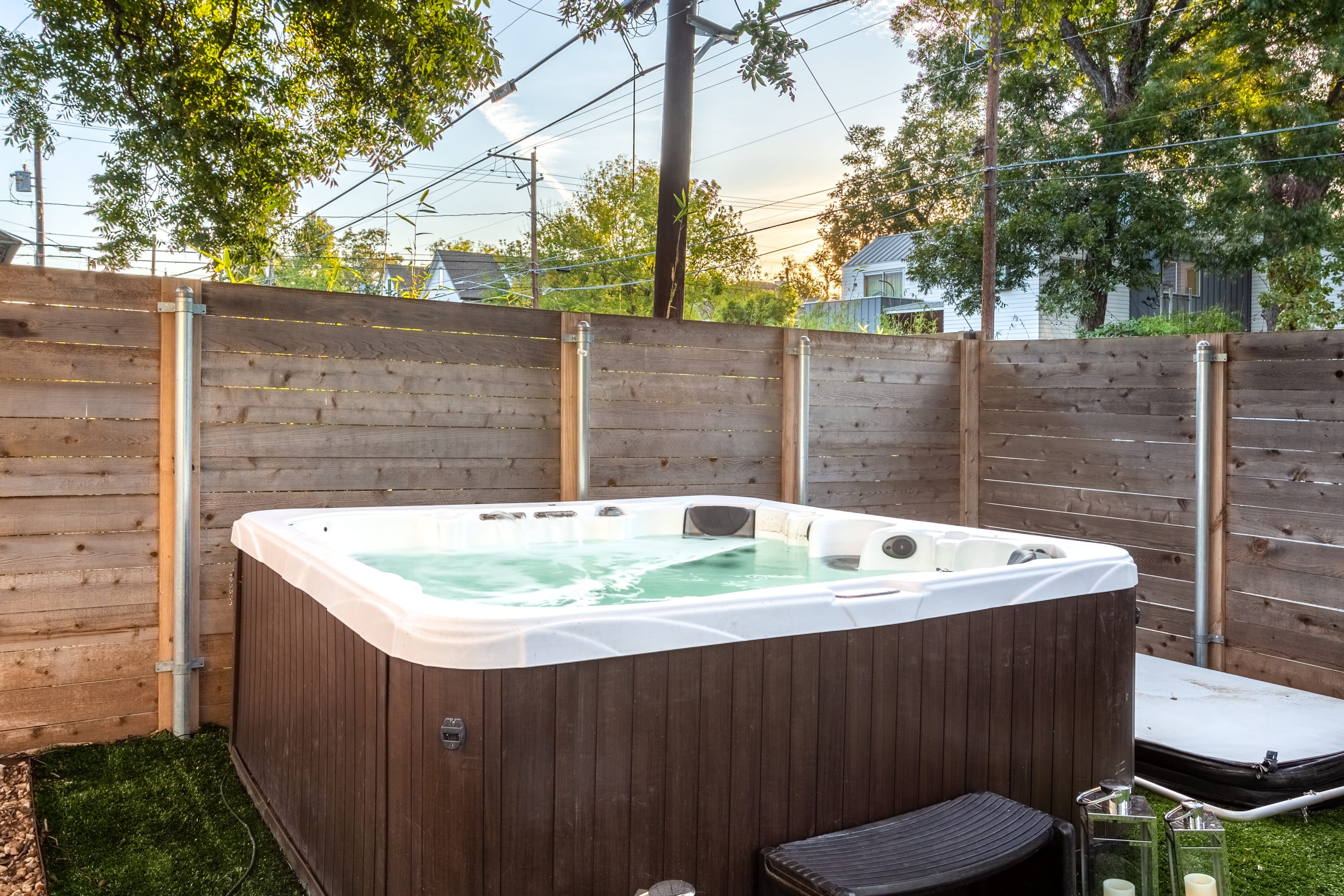 Close-up view of a bubbling hot tub with a dark brown exterior and white interior, set on artificial turf within a fenced backyard. The water is visibly agitated, indicating it's in use.