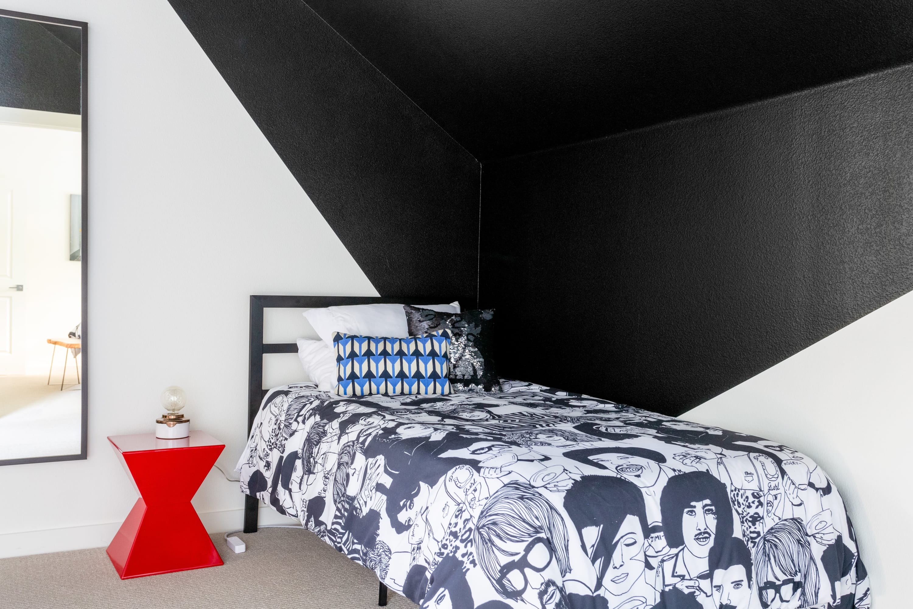 Modern bedroom with bold black and white geometric walls, featuring a single bed with a black and white patterned duvet and a red side table with a lamp.