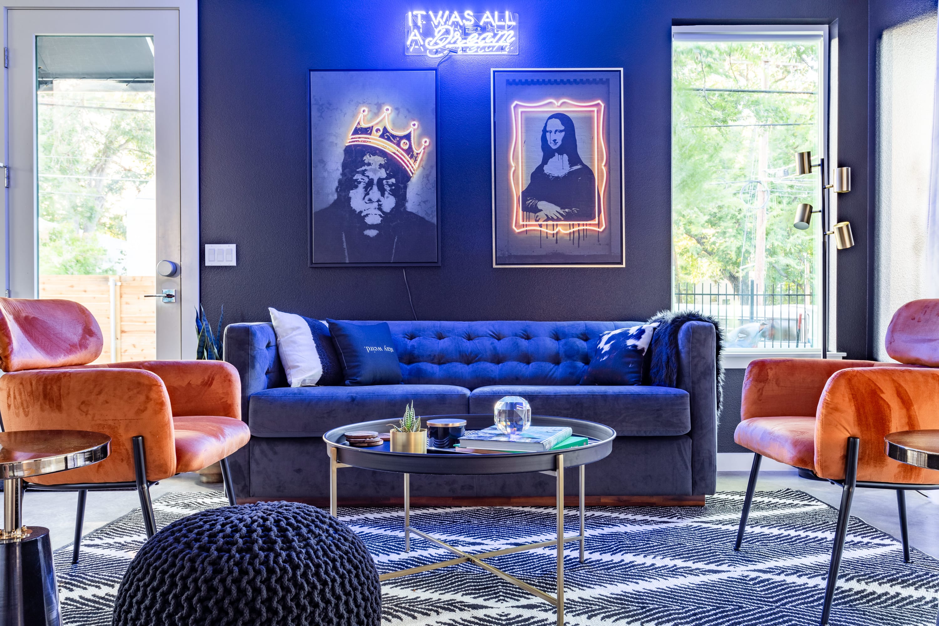 A vibrant living room features a dark blue tufted sofa, two orange velvet armchairs, and a round coffee table on a black and white geometric rug, with neon art on a dark wall.