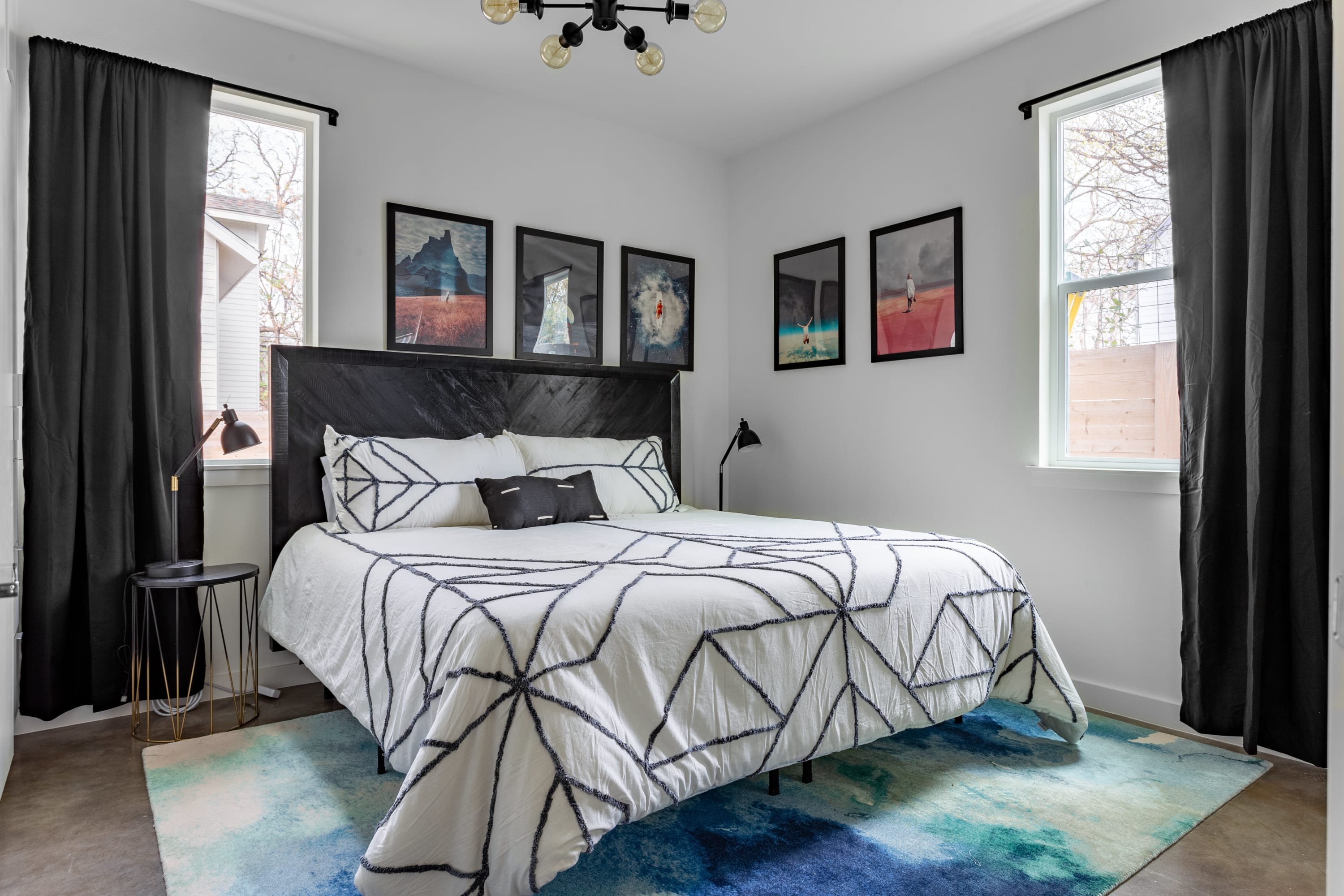 Spacious bedroom featuring a king-sized bed with white bedding adorned with black geometric embroidery, a dark wooden headboard, and black room-darkening curtains. Multiple framed art pieces hang above the bed, and a vibrant blue and green area rug covers the floor.