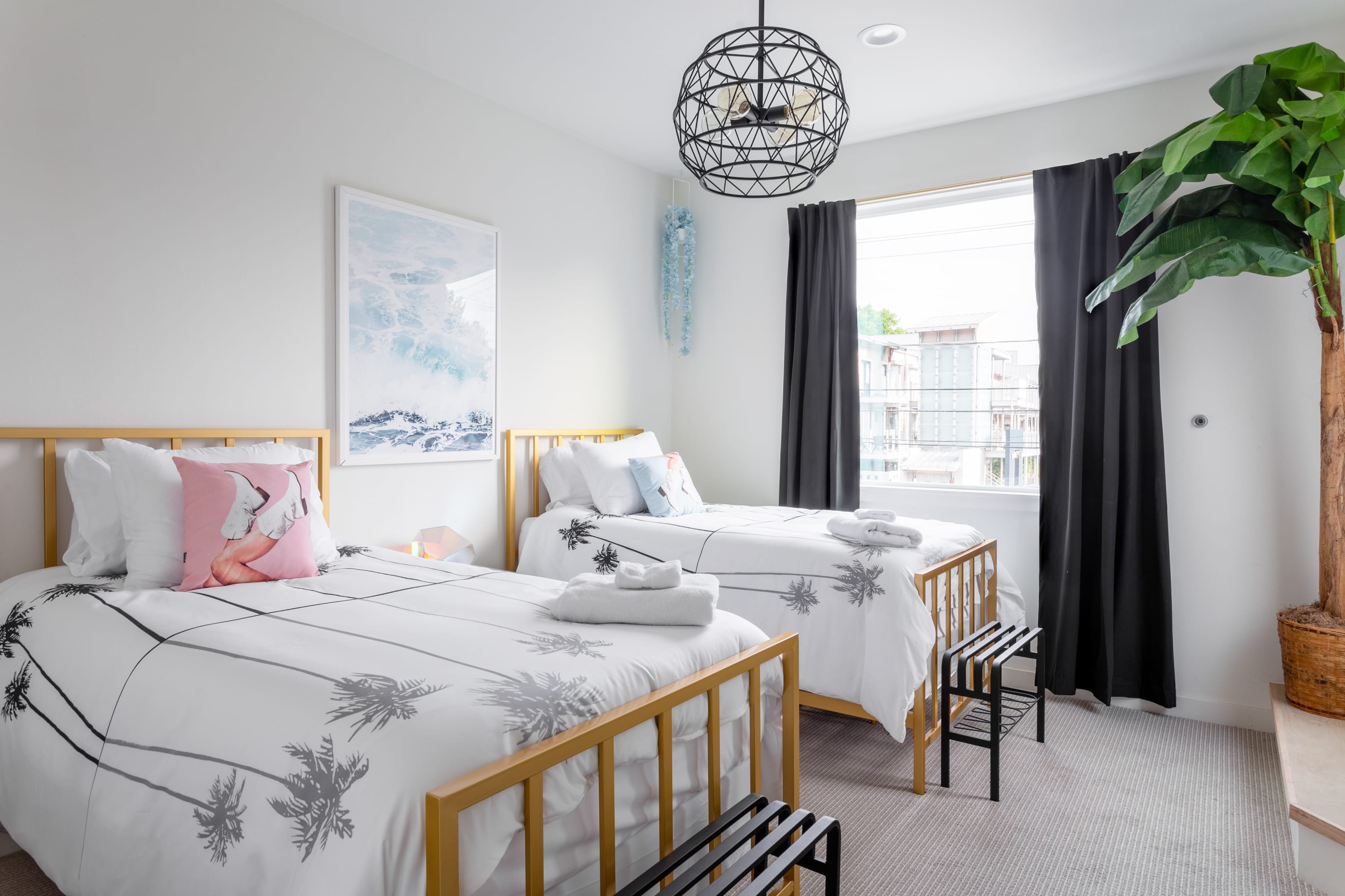 A bright bedroom features two gold-framed twin beds with white bedding adorned with palm tree patterns, a pink flamingo pillow, and a blue ocean art print on the wall. A black geometric chandelier hangs from the ceiling, and dark curtains frame a large window.